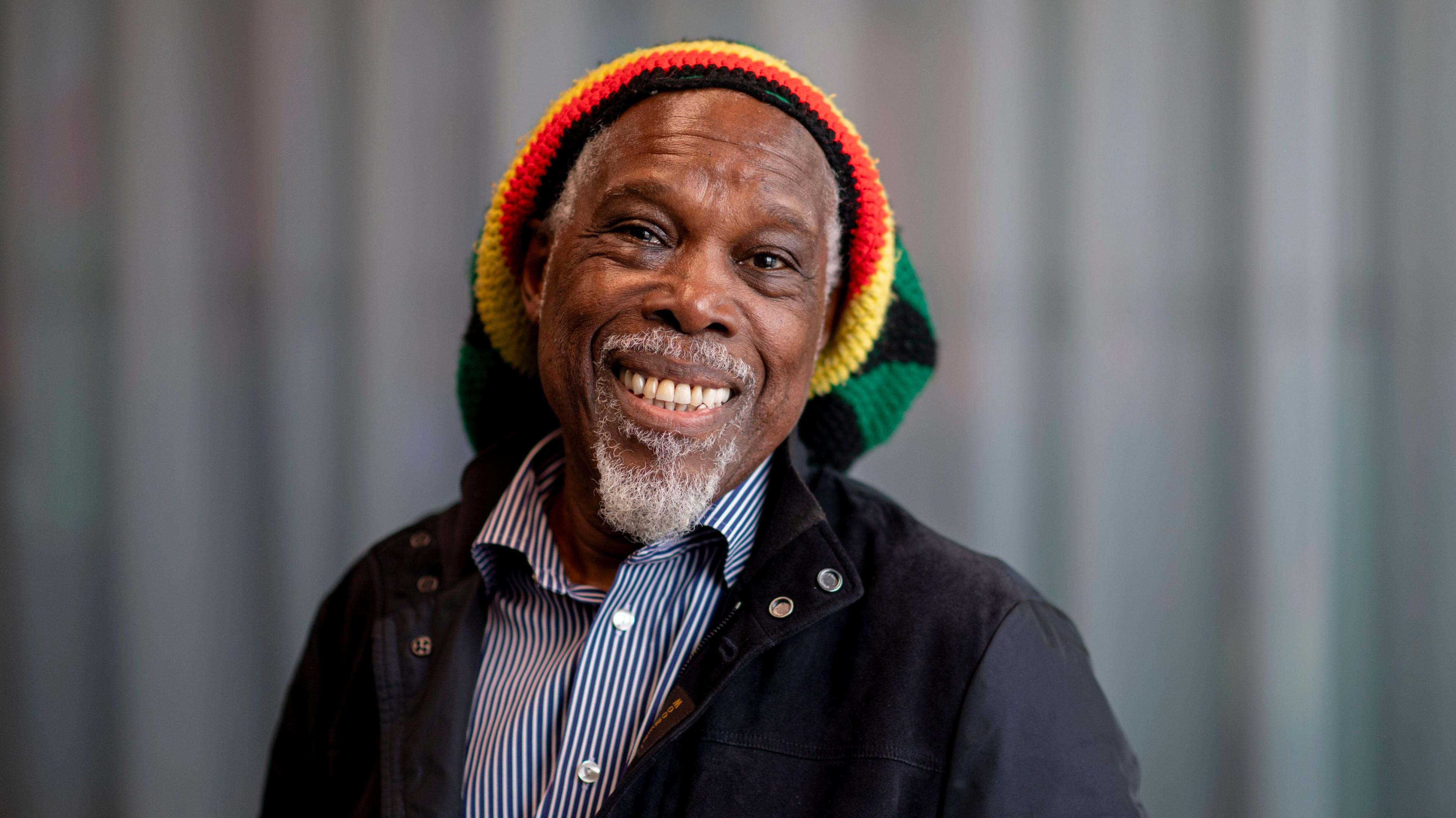 A man (Billy Ocean) is wearing a red, yellow, green and black striped hat and smiling into the camera. He has a short white beard and moustache. He is wearing a blue and white striped shirt and a black jacket. A blurred grey background is seen behind him.