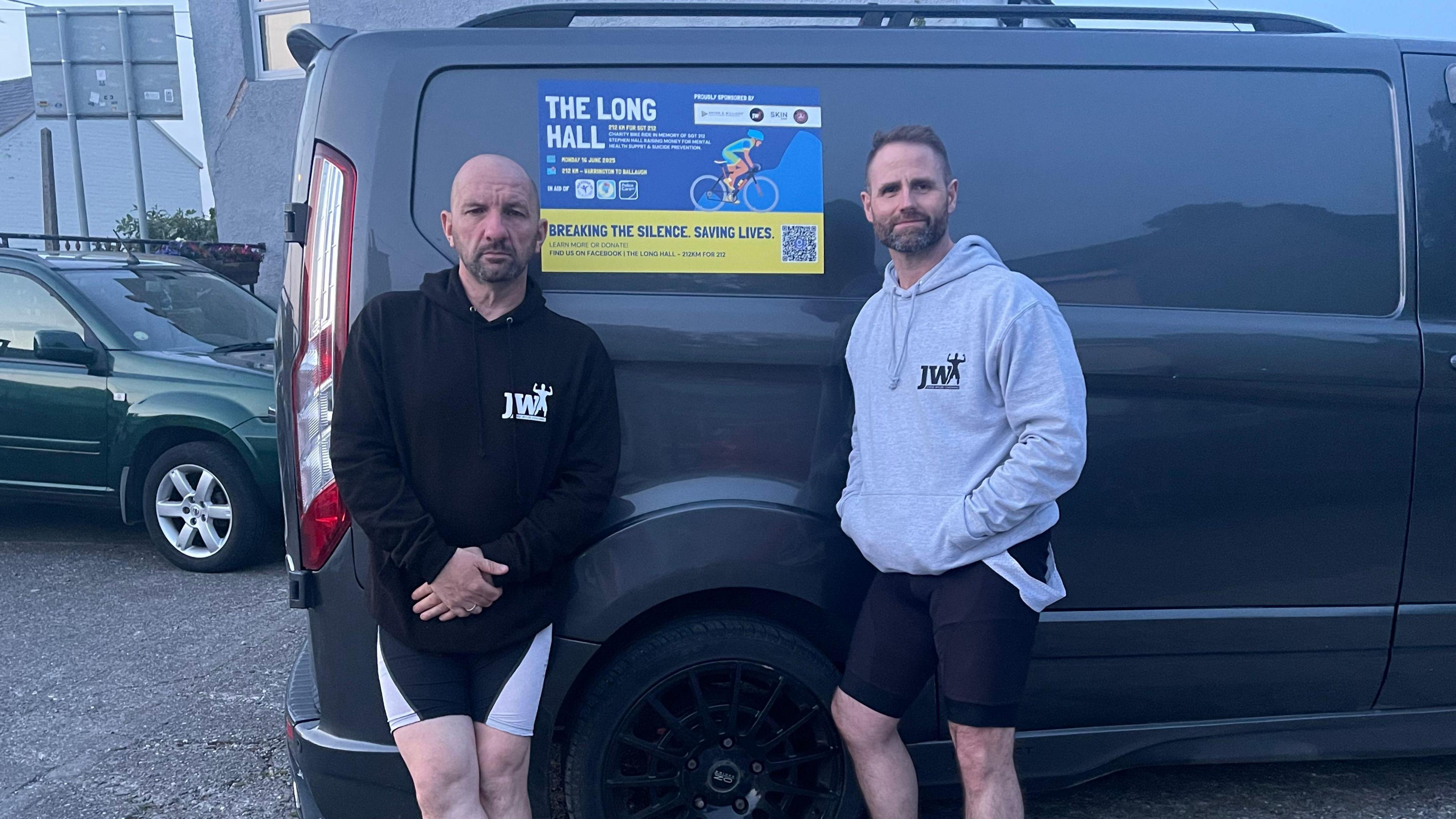 Adrian a bald man in a black jumper and Darren a man with brown hair and stubble wearing a grey jumper stand next to a black van with a poster that says the long hall.