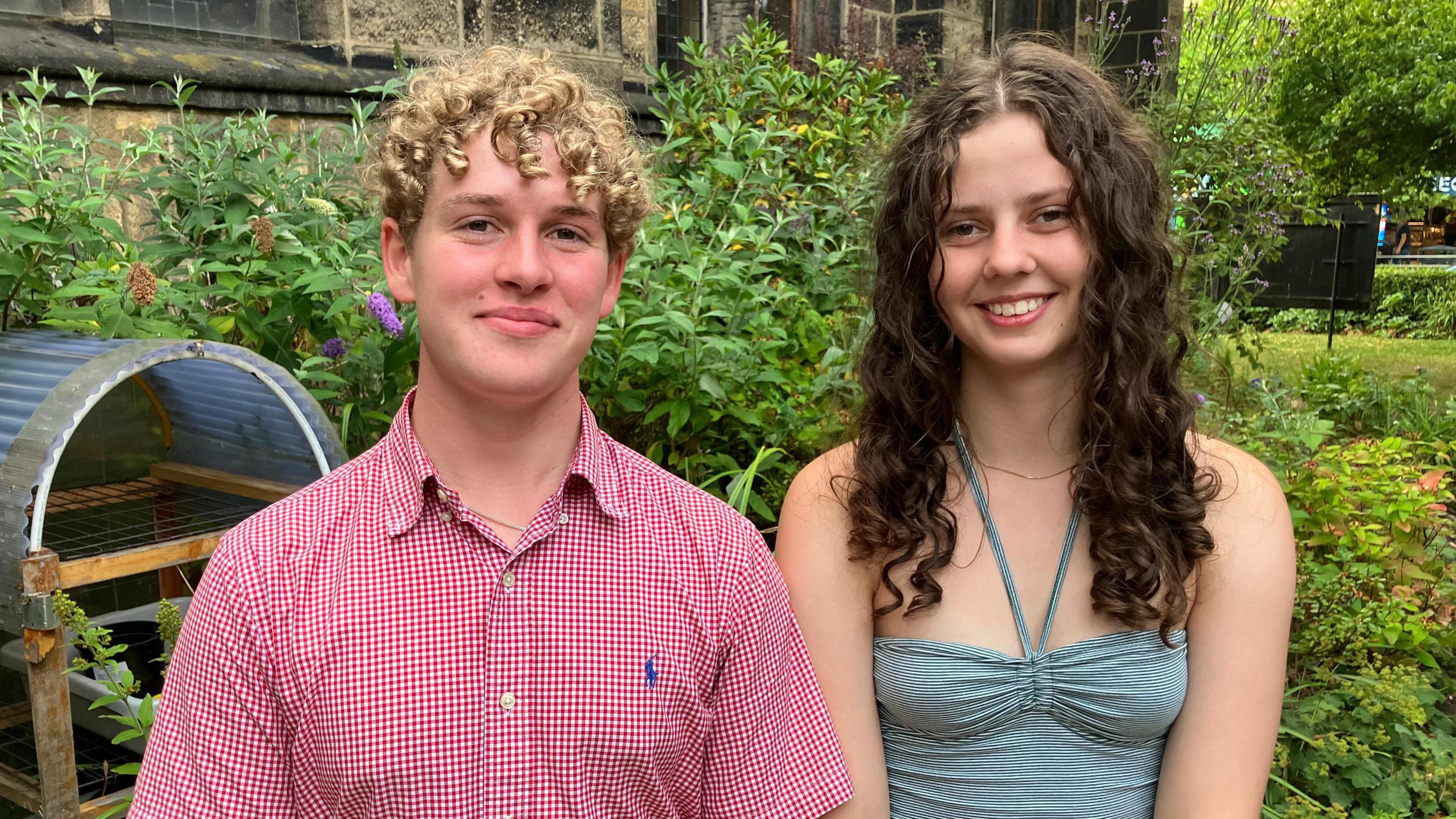 A young man and young woman smile at the camera. They are standing in a garden, with plants behind them.