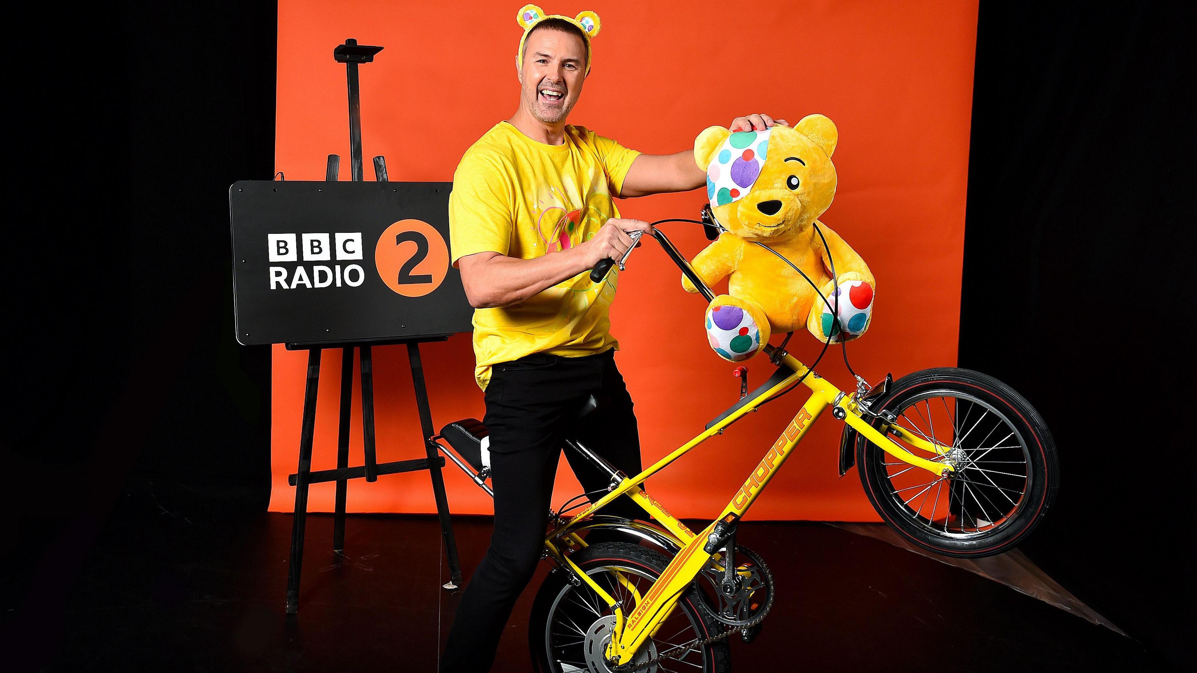 Paddy McGuinness stands holding a yellow bike which is balanced on one wheel and a yellow Pudsey bear mascot. Paddy is wearing yellow bear ears and a yellow t-shirt. He is standing in front of a bright orange backdrop and there is a sign that says "BBC Radio 2"