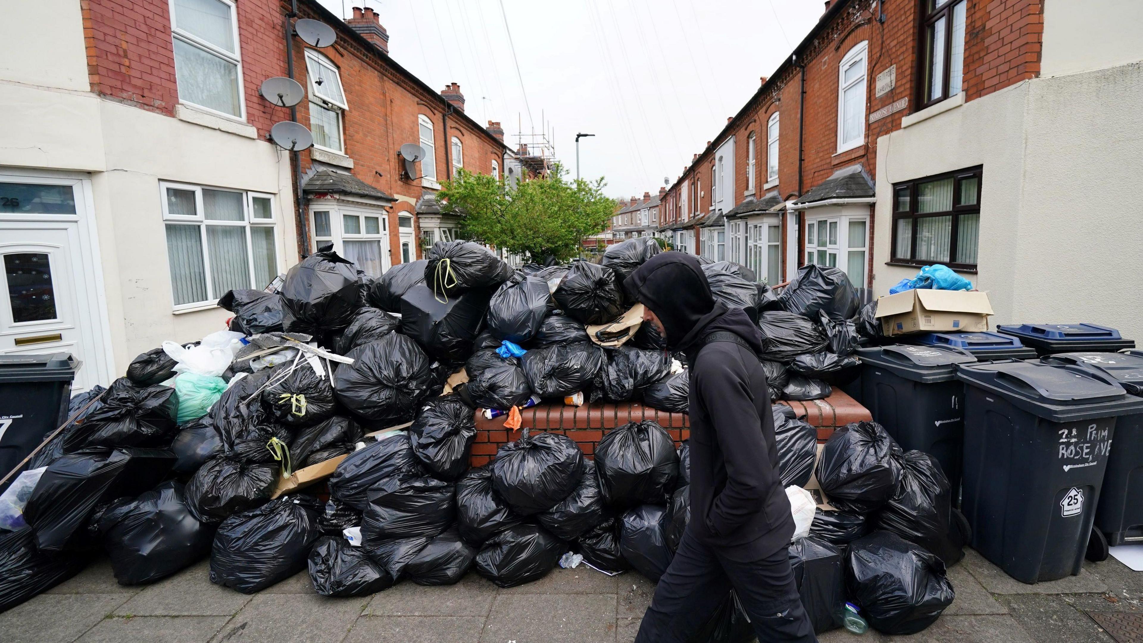 A man in a black hoodie walks past black bin bags piled up on a residential street, with terraced houses on either side.