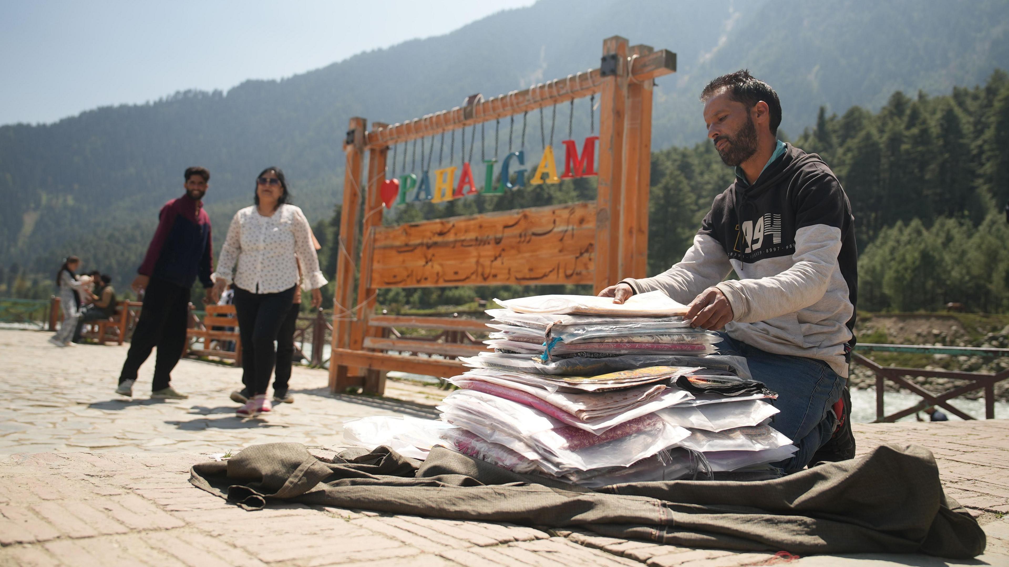 The image shows a shawl seller sitting next to a tourist board that says "I love Pahalgam" and a few tourists behind him.