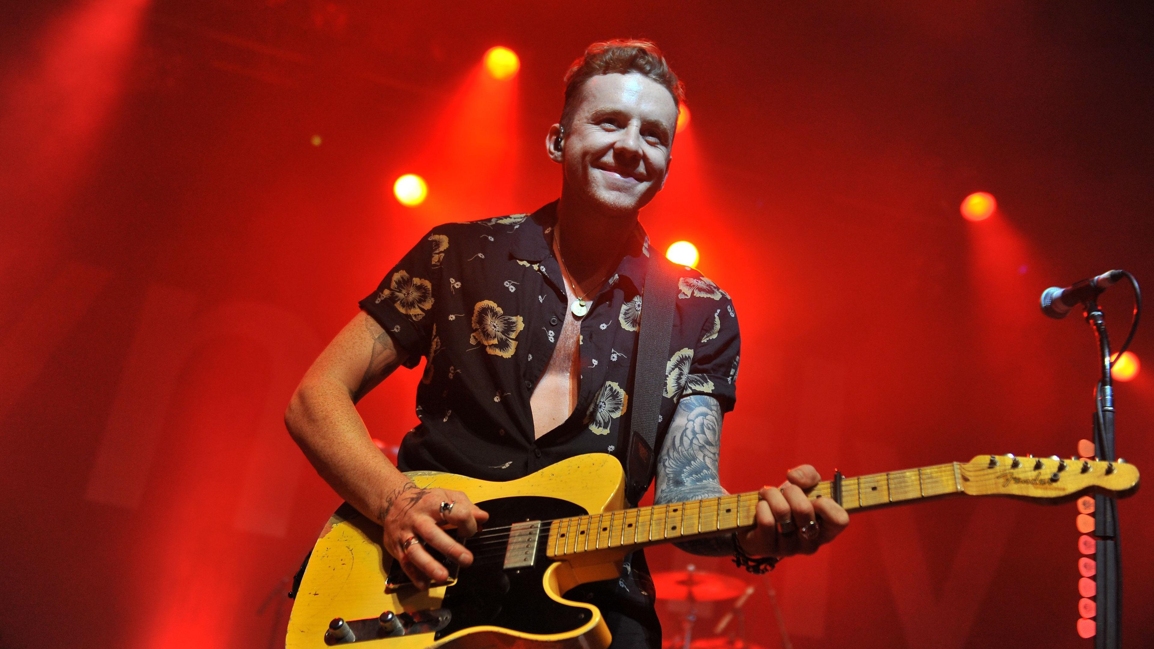 Danny Jones smiling while playing an electric guitar on stage. He is wearing a black flowery shirt. Red lights brighten up the background behind him