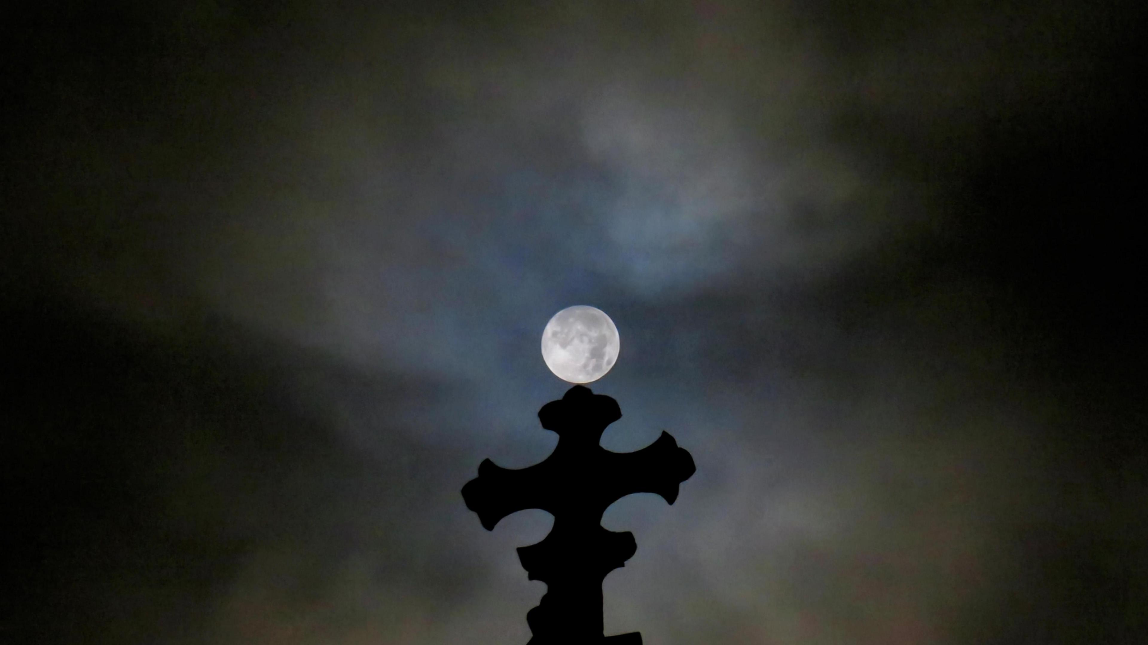 Full Moon seemingly perched on top of a church cross, in a dark sky with thin cloud