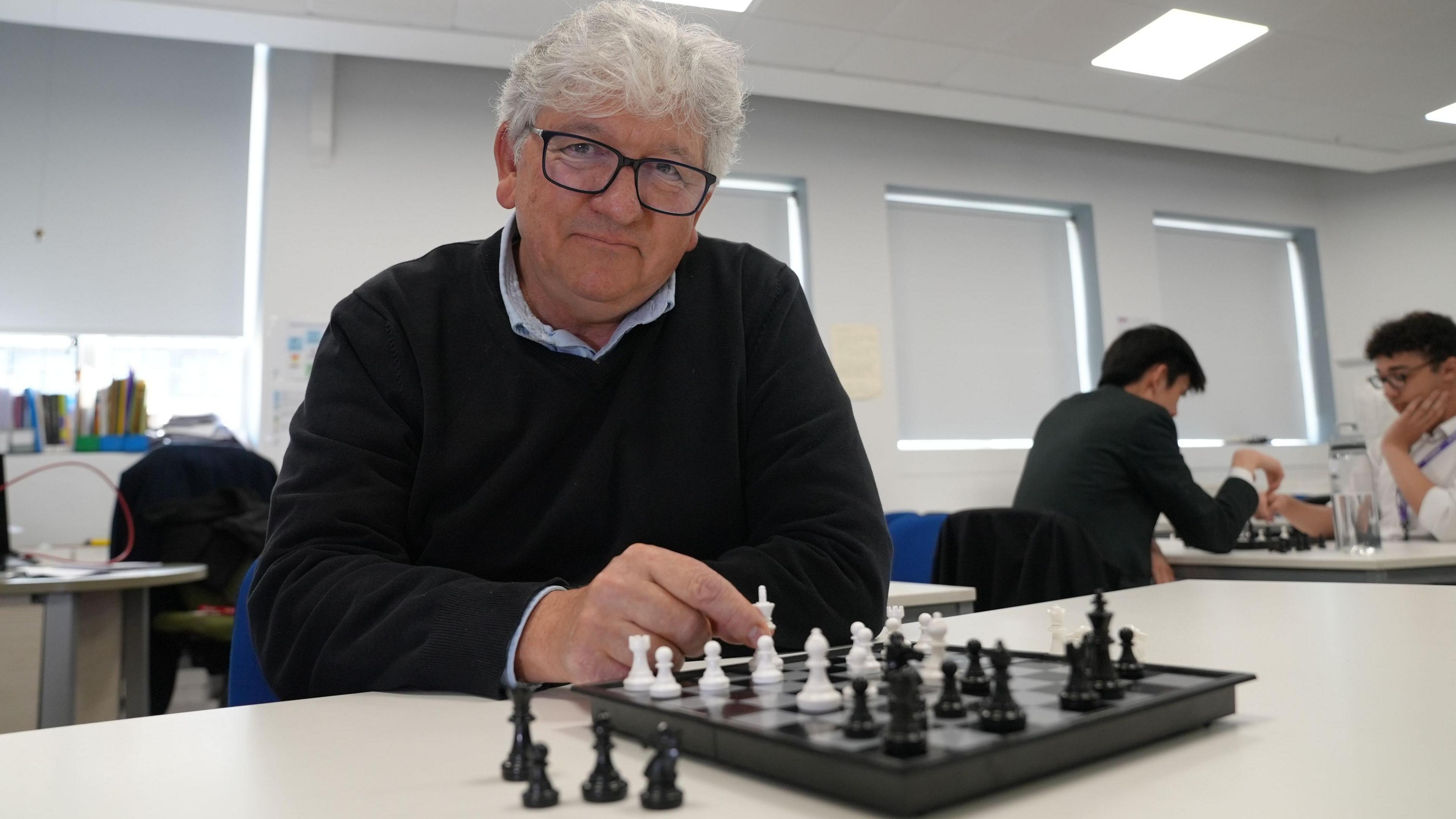 Andrew Maffessanti is wearing a black jumper and is holding a white king on the chess board in front of him. He has white hair and black glasses and is smiling. Two students can be seen in the background playing chess too.