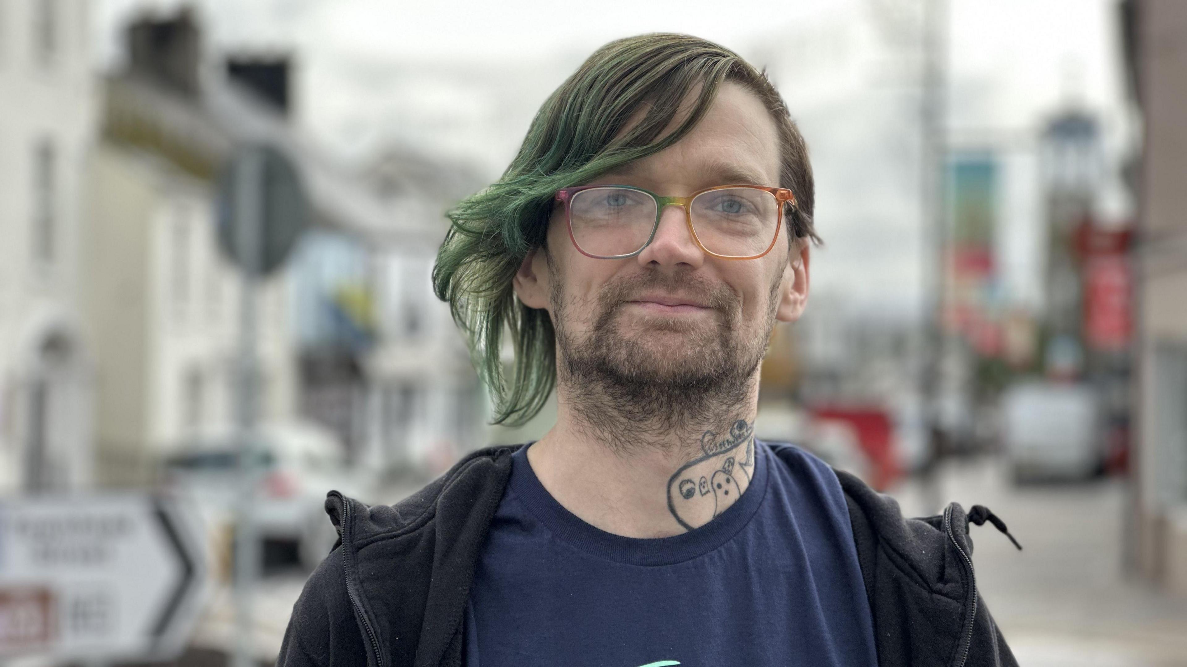 Riley has green tinted hair. He is wearing glasses and has stubble. Riley is wearing a navy top and a black zip up jacket. He has a tattoo on his neck. He is smiling. The street behind him is out of focus.
