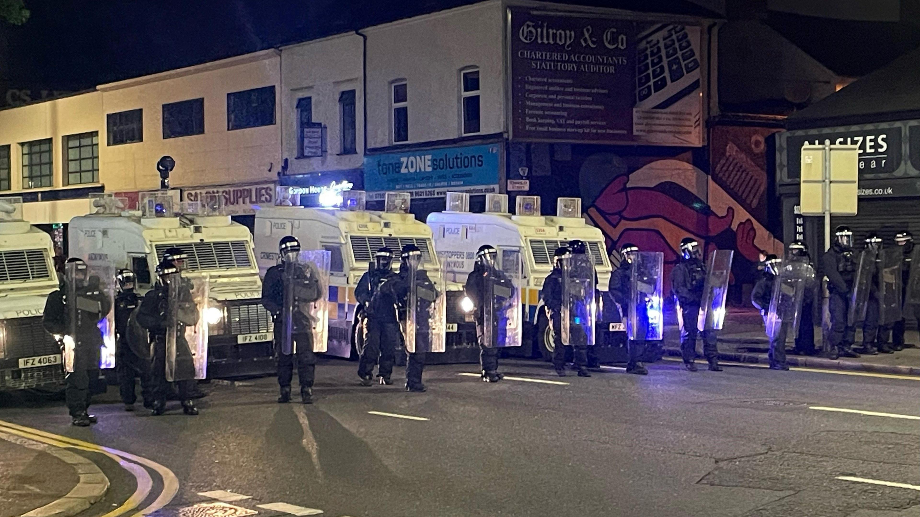 A line of police land rovers with police in riot gear in a line standing in front of them in east Belfast.