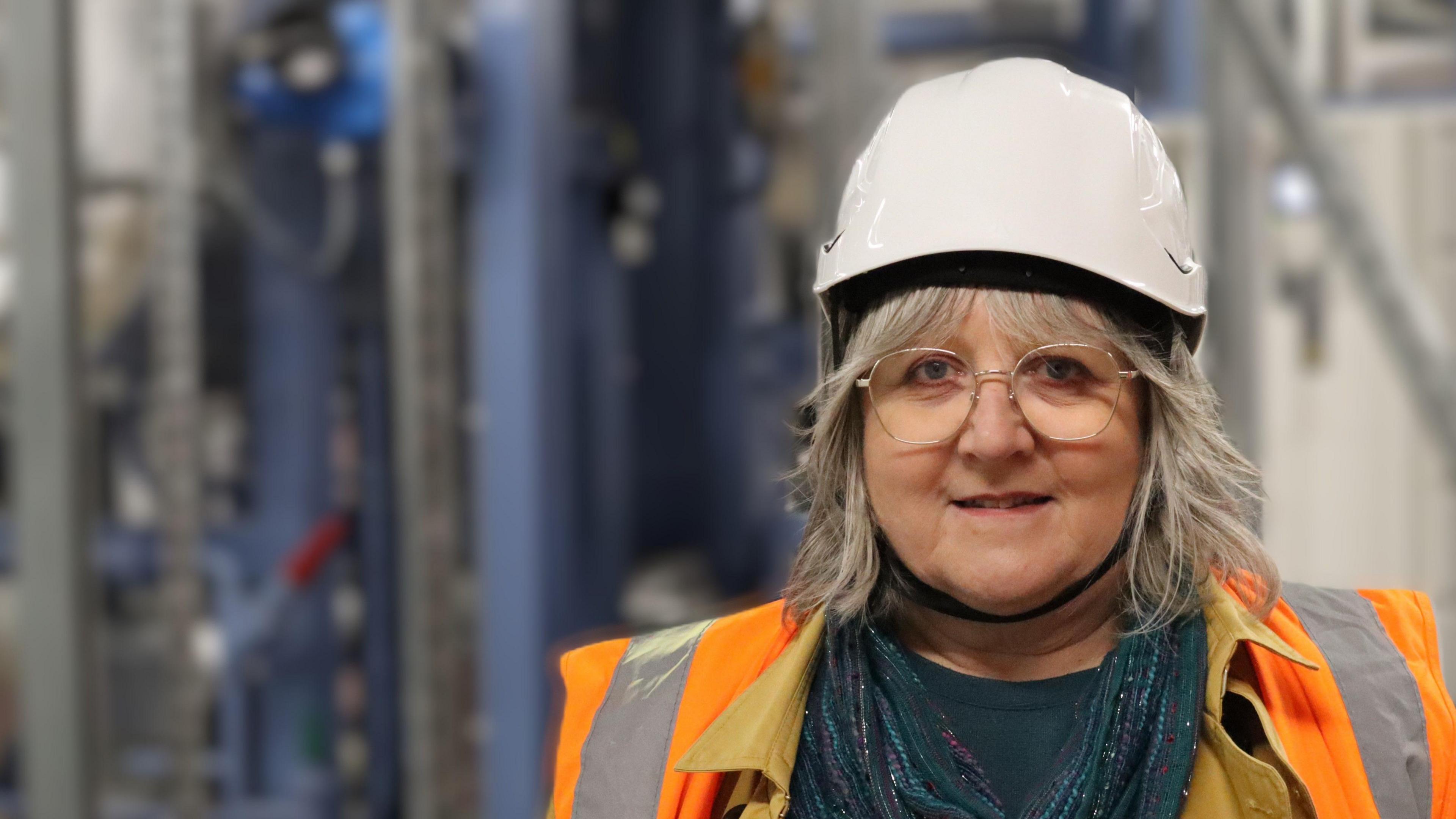 Lorna Dupre wearing a white hard hat and orange safety vest, looking at the camera. She has glasses, long grey hair and is wearing a teal pashmina.