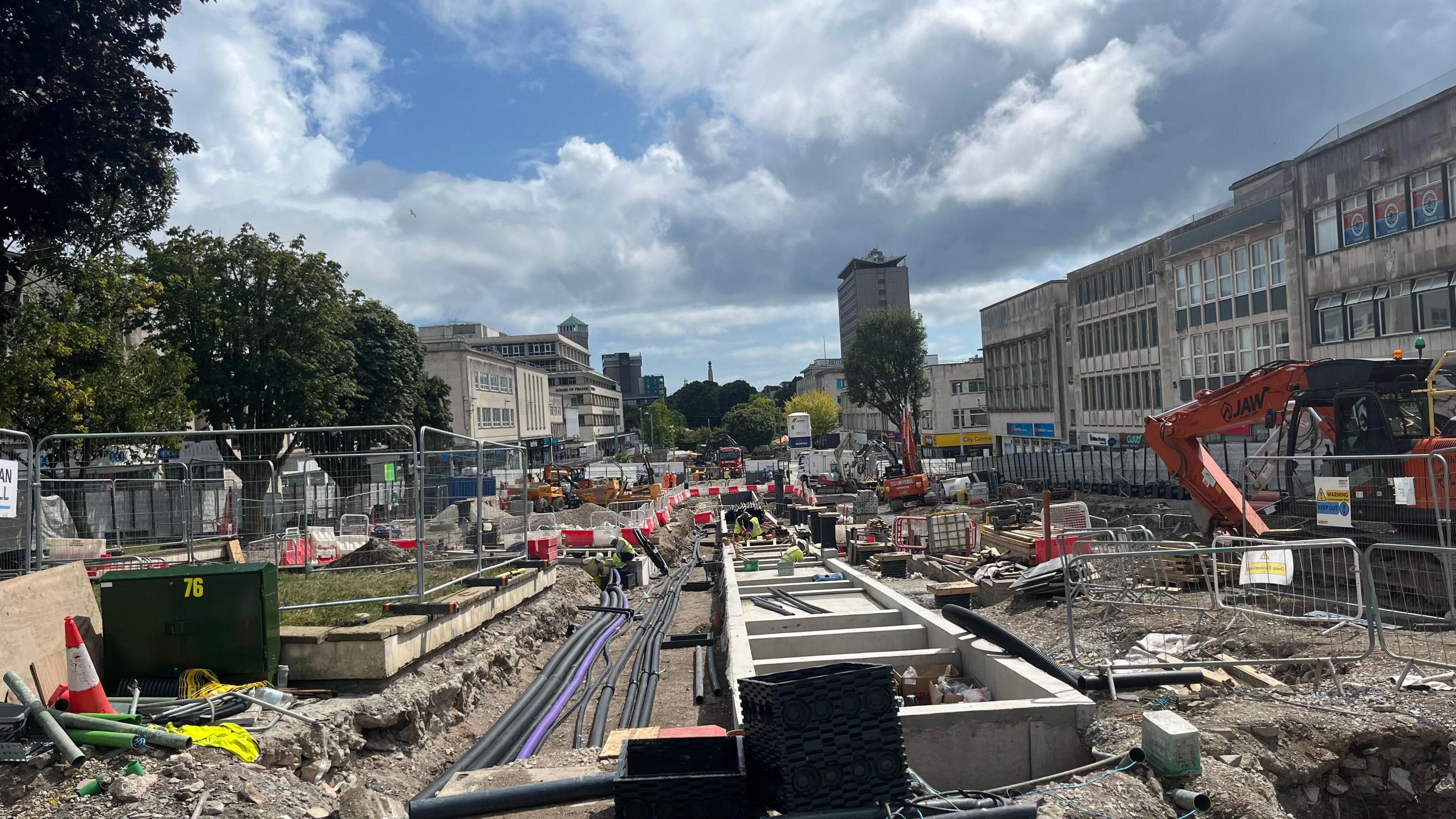 Plymouth Armada Way being dug up. There are diggers and soil. There are shops on either side. The image is looking down towards the sundial. There are trees to the left.