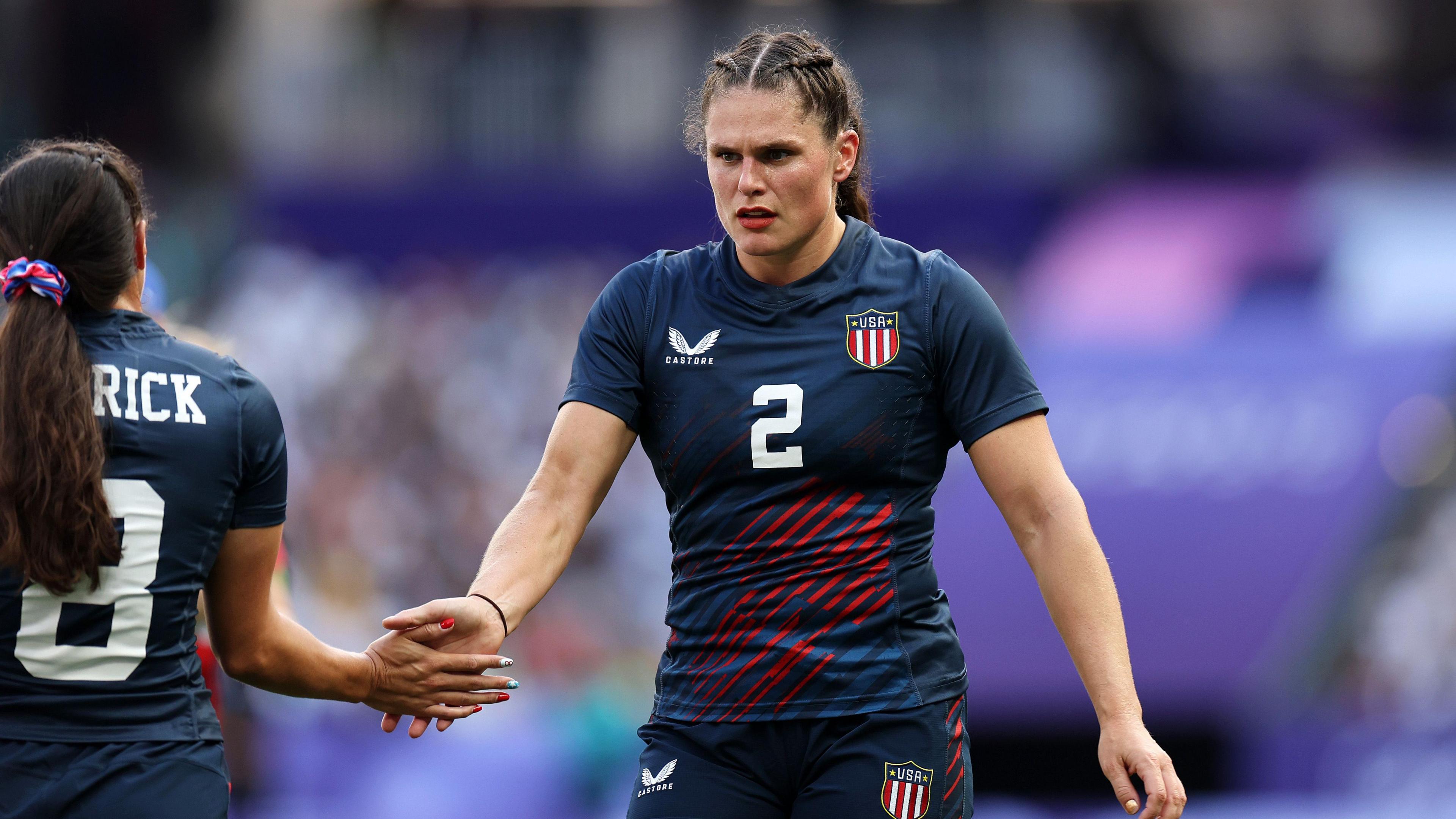 Two women rugby players. The one on the left has her back turned with long brown hair in a ponytail. She is high-fiving the other rugby player who is facing the camera. She is wearing a blue, number 2 rugby top and has her hair in two plaits.