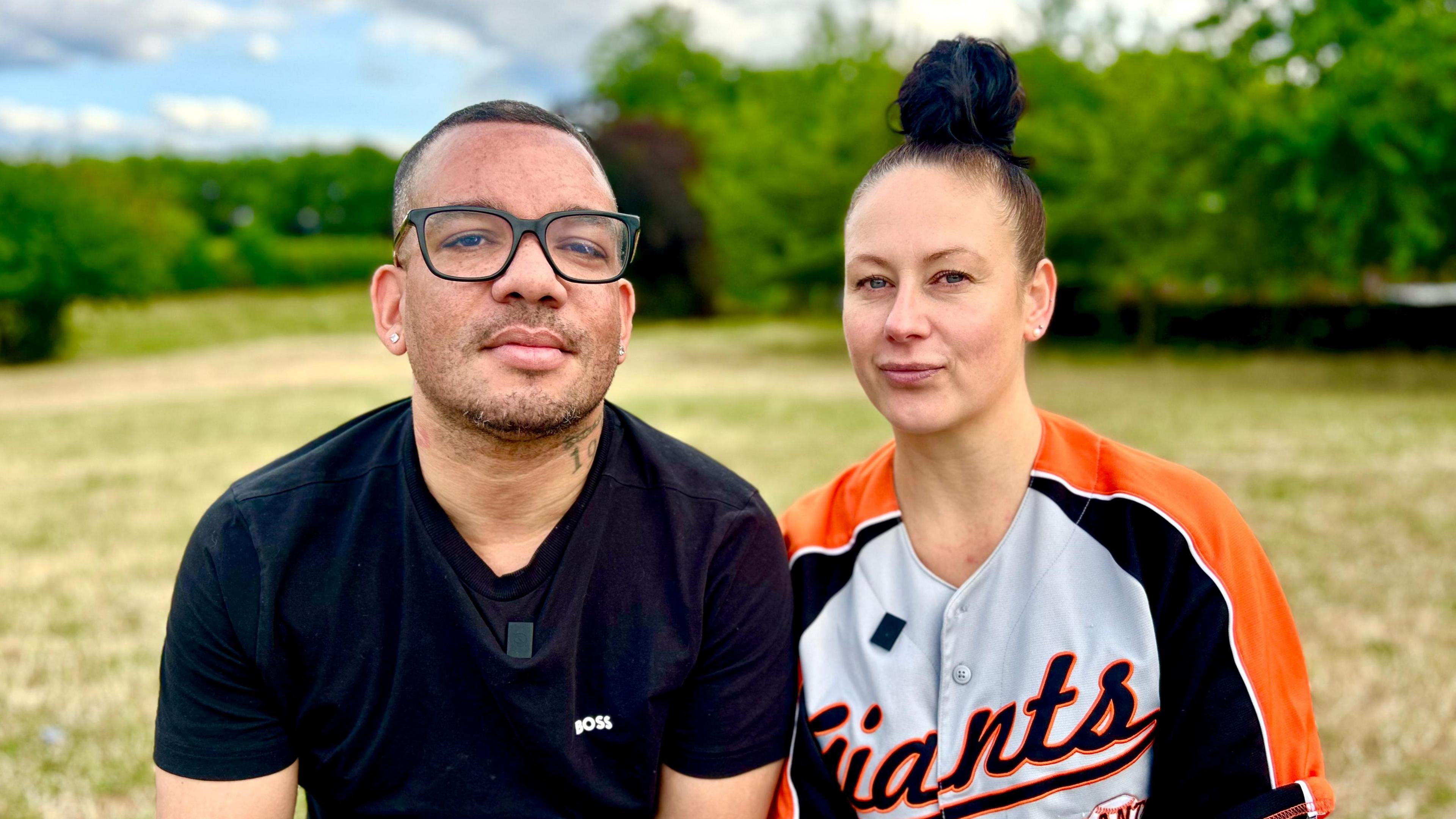 Kester and Zoe are sitting outside in a park looking directly at the camera. Kester is wearing a black tee shirt and thick, dark-rimmed glasses, and has a small tattoo on the left of his neck. Zoe has her brown hair tied in back and up in a bob and has a wry, closed-mouth, smile. She is wearing an orange, black and light grey baseball top.