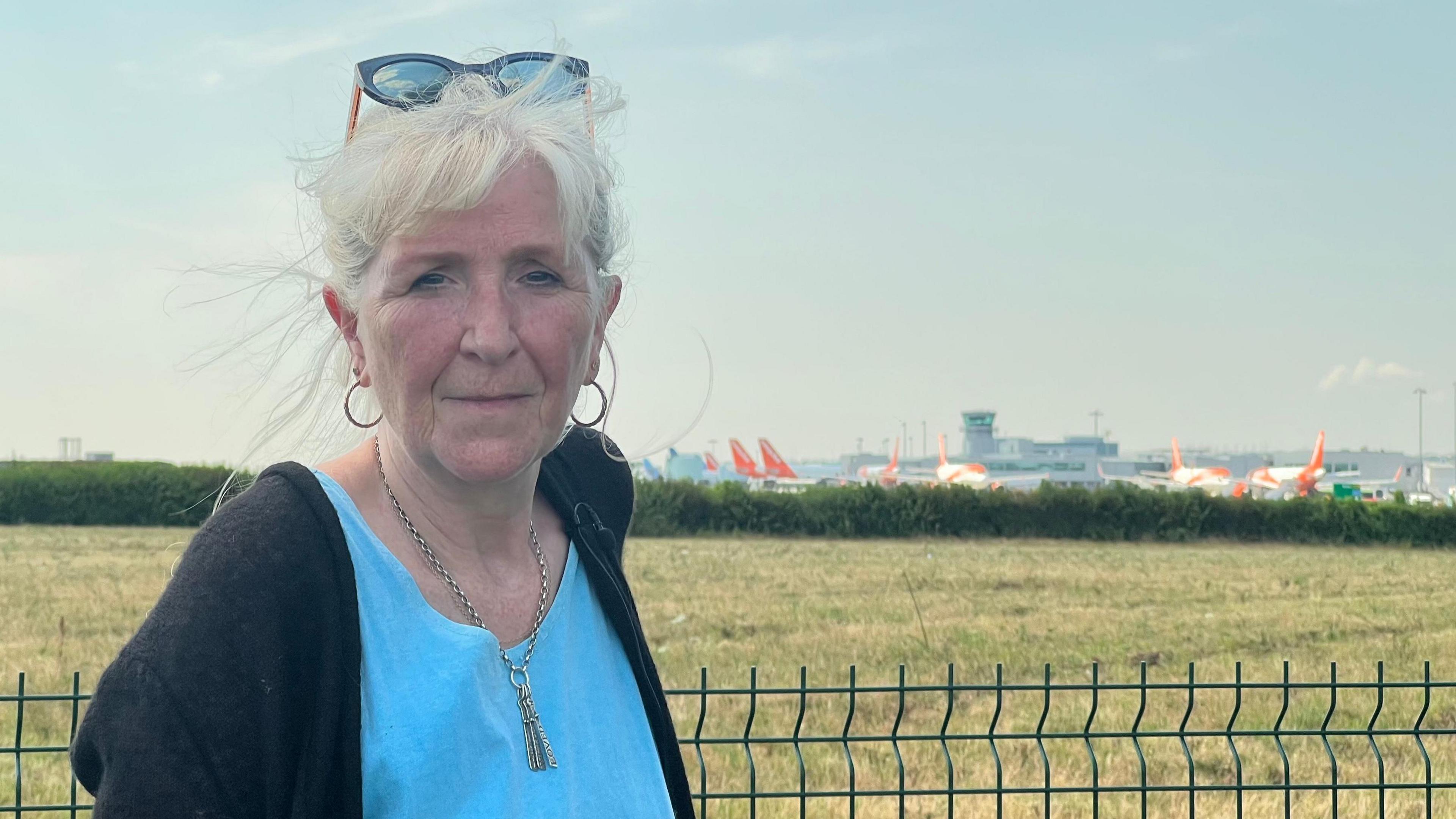 Mary Collett, a woman with white hair and sunglasses on the top of her head, is standing in front of a green field with Bristol Airport on the horizon.