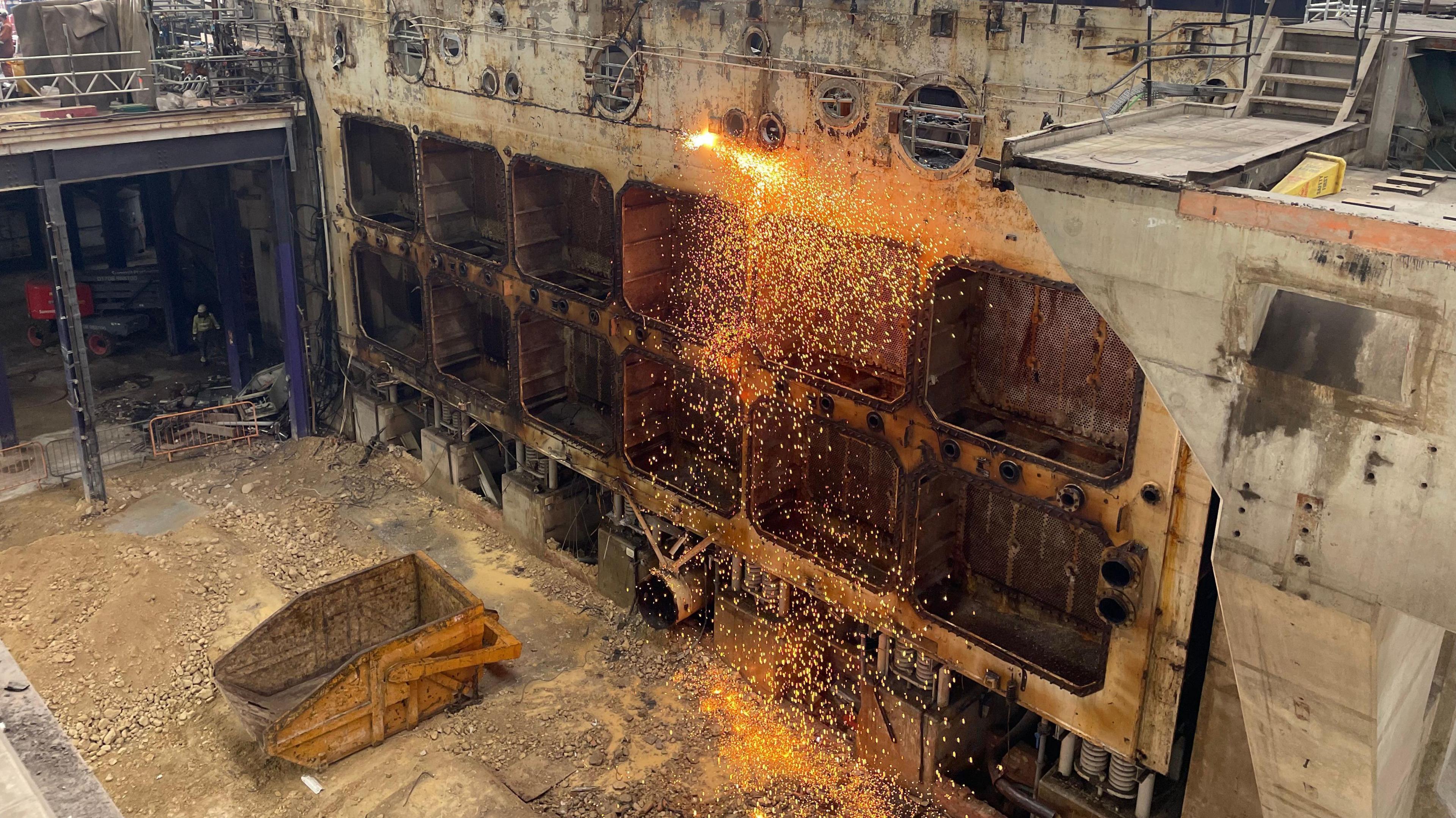 Sparks flying off a wall inside the turbine hall. There is a yellow skip on the ground, with various platforms also pictured in the hall.