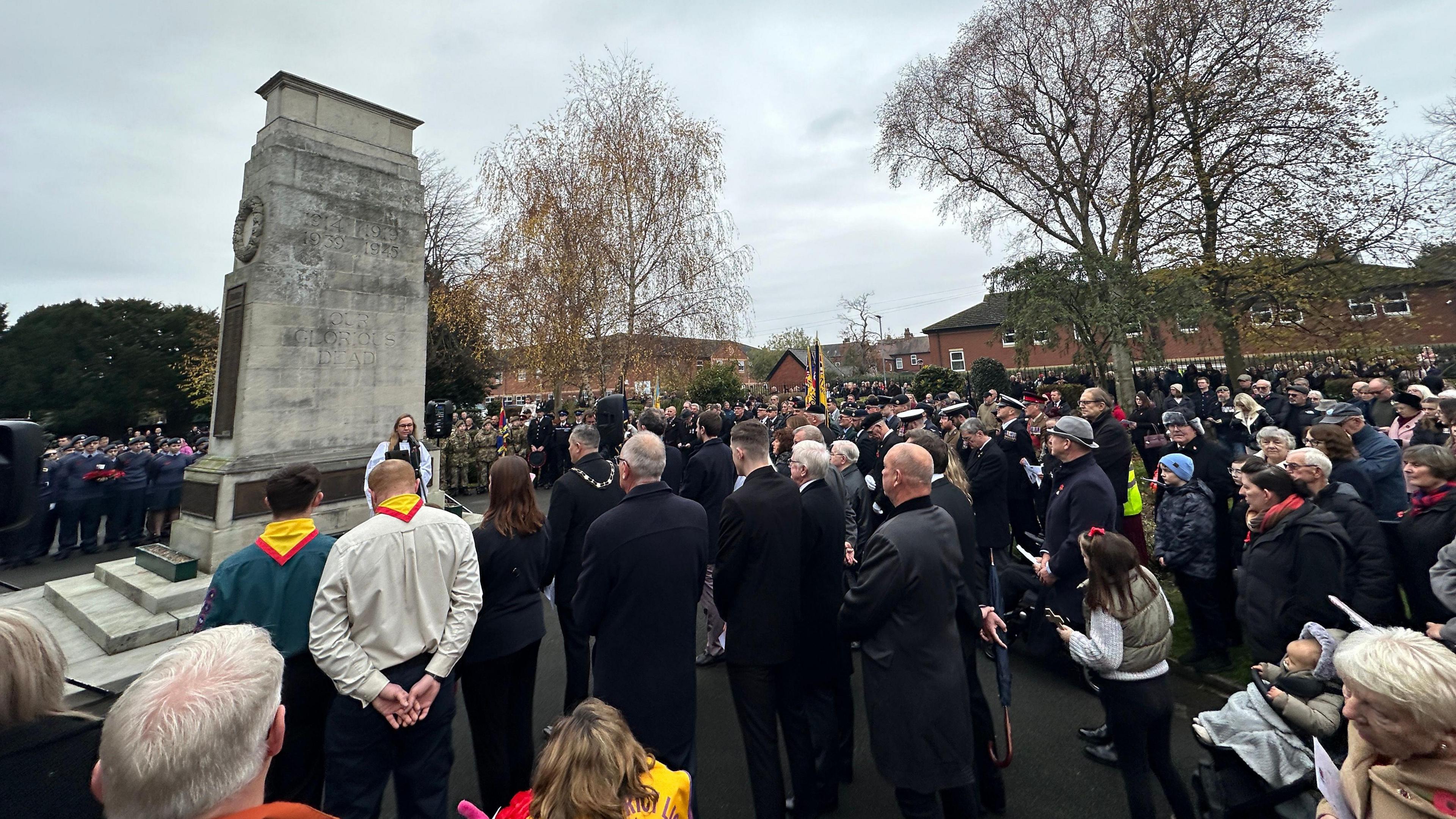 Crowds gather at the cenotaph, most in black with their heads bowed.