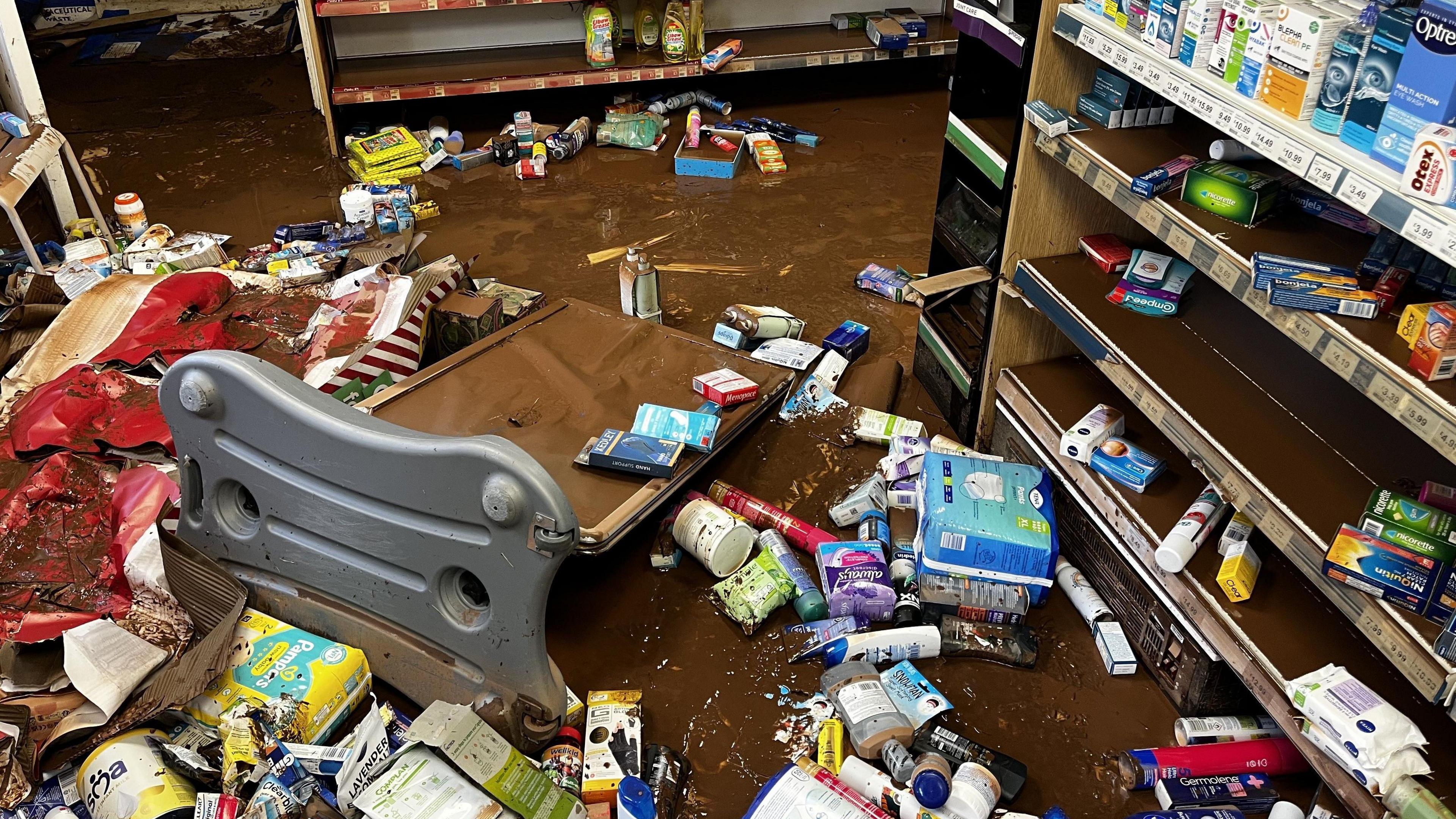 The floor of a flooded shop with medicine, toiletries and packs of baby food and nappies floating in the water.