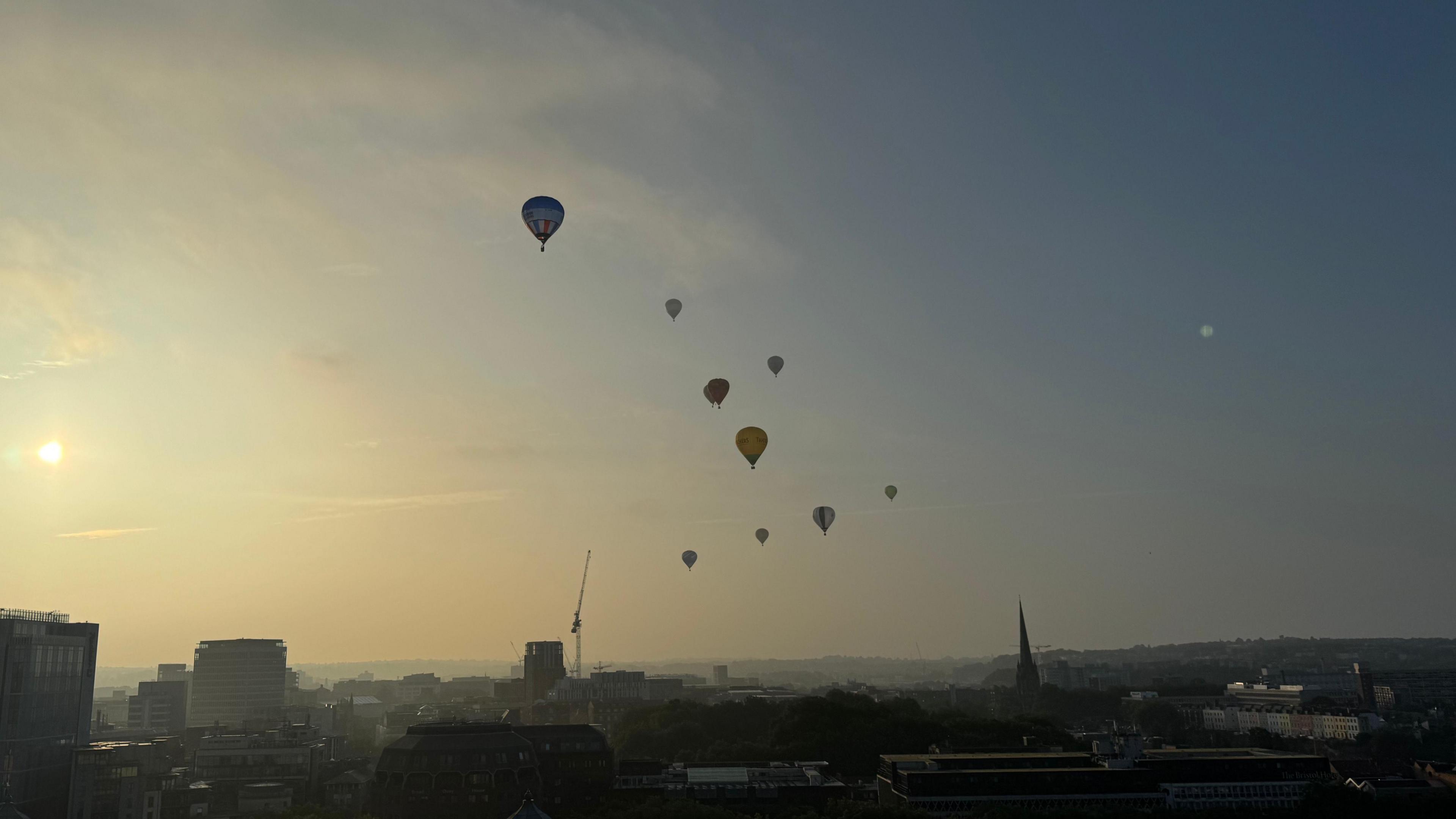 Nine hot air balloons flying above Bristol in the distance. The city skyline, which features high rise buildings, a church spire and cranes, can be seen. 