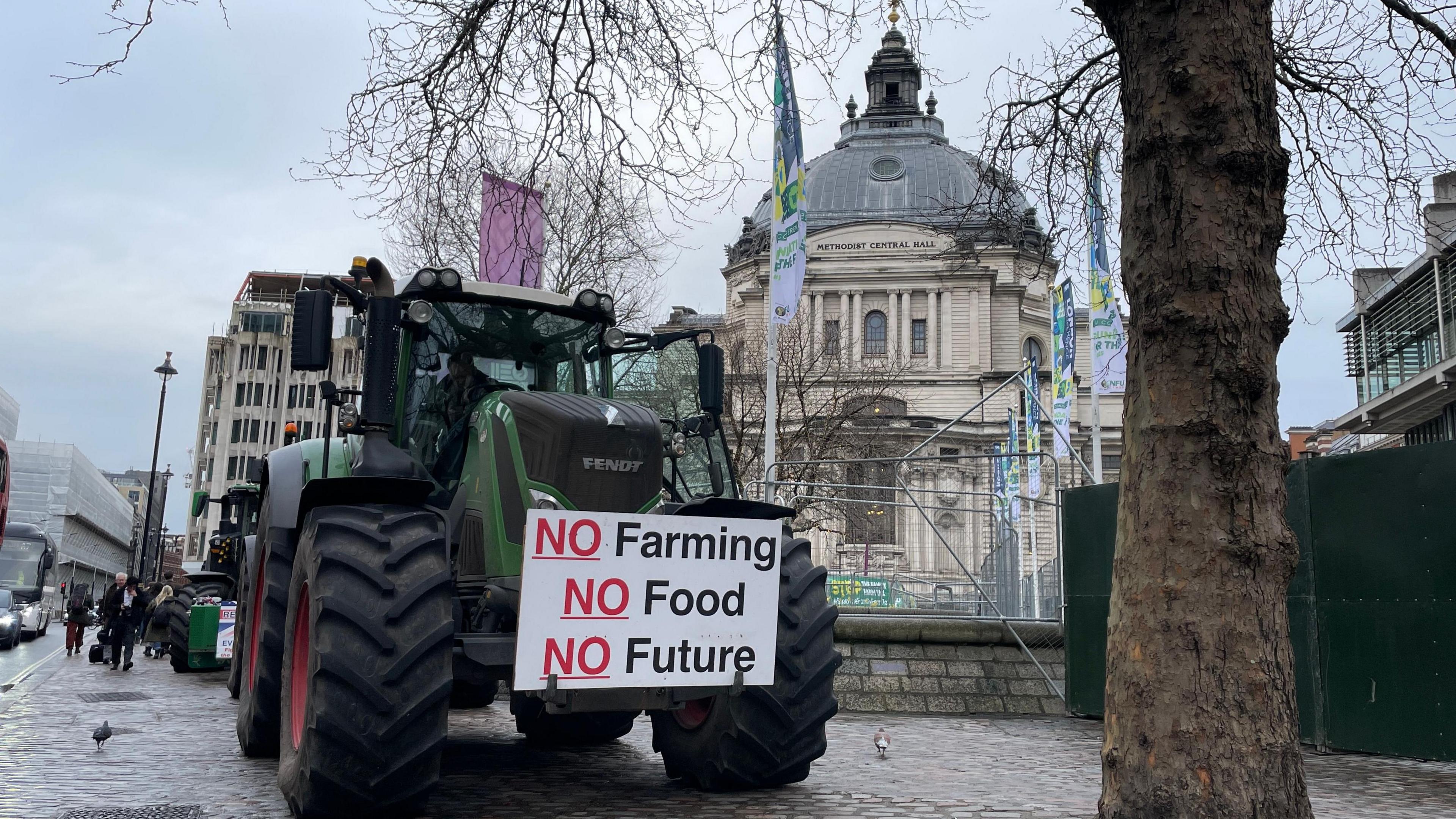 A green tractor is parked on a Westminster pavement under a grey winter sky next to a bare tree and in front of the QEII Centre. On the front of the tractor is a protest placard that reads No Farming, No Food, No Future. Pedestrians pass by.
