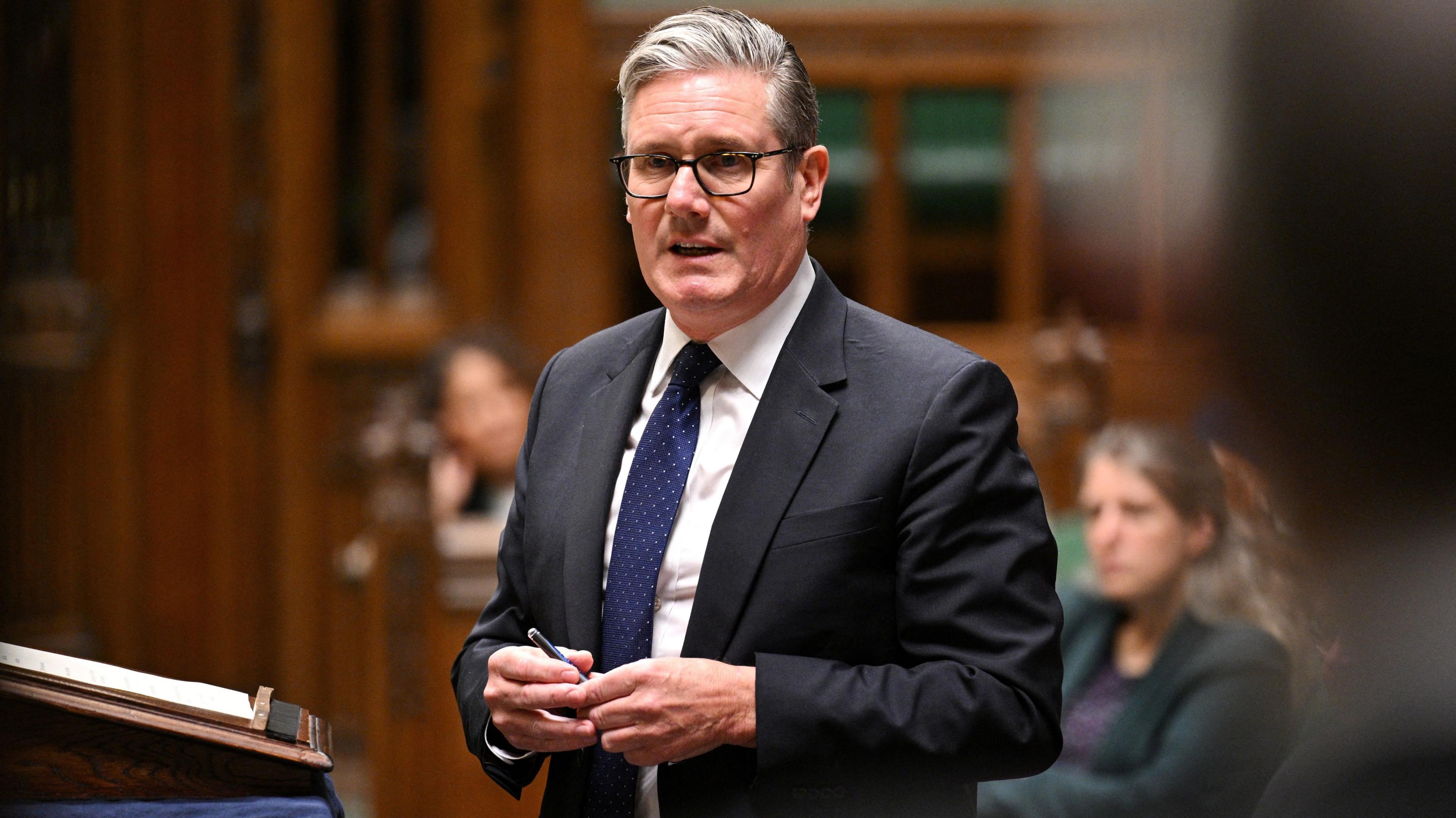Sir Keir Starmer stands and speaks. He wears a black suit, white shirt and blue tie.