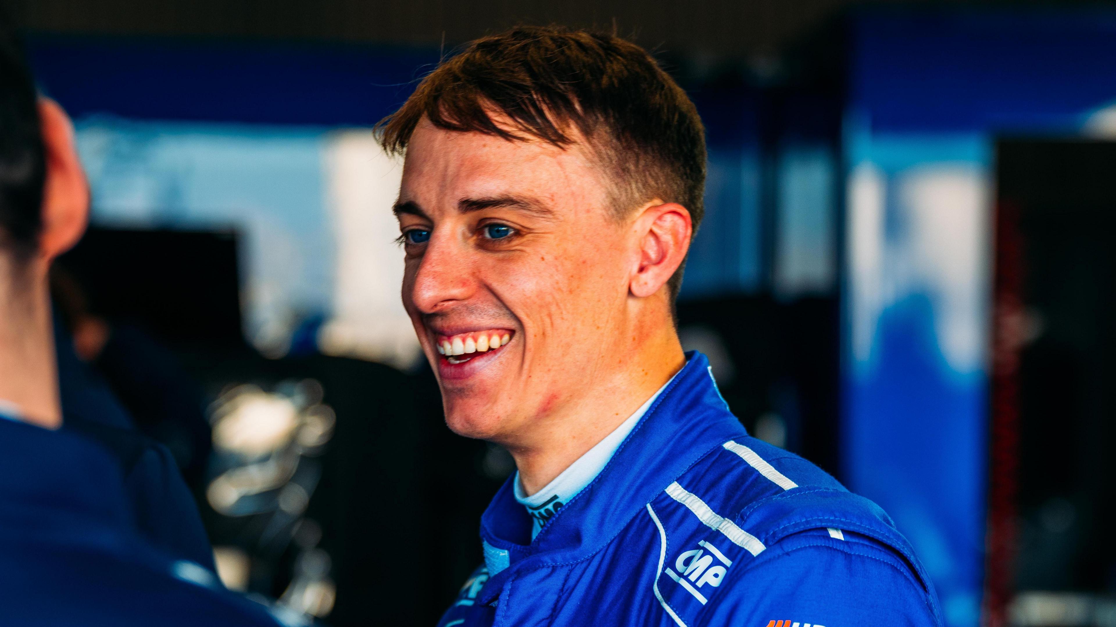 A man with short brown hair and wearing a blue racing suit smiles as he looks to the left at someone.