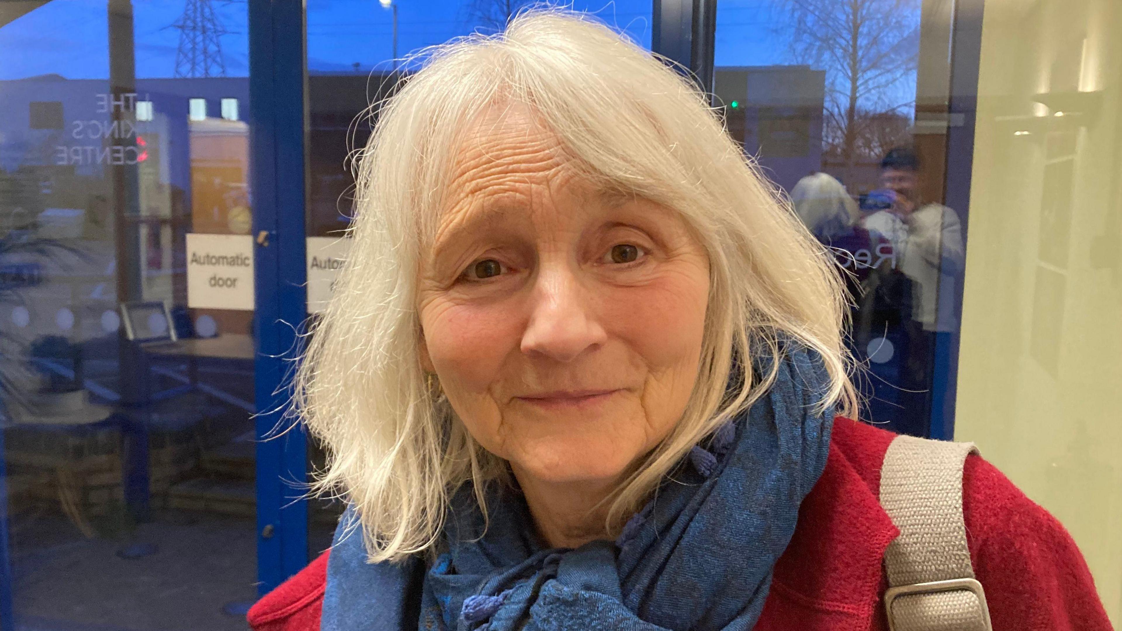 Sue Spencer-Longhurst is wearing a red coat and a blue scarf in the King's Centre foyer and there are blue double doors behind her. 