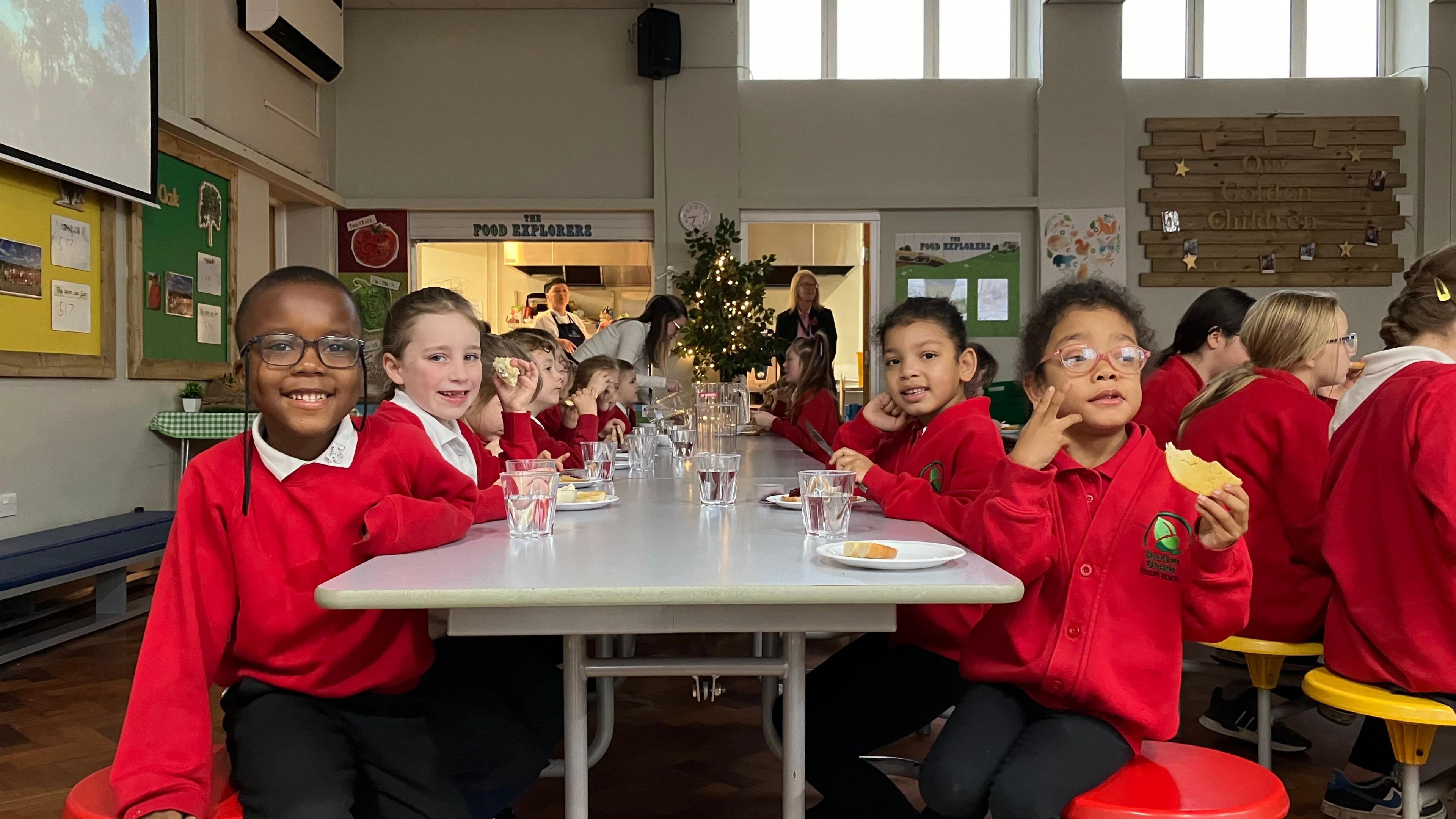 A group of primary school children dressed in red uniforms, sit on either side of a long dining table eating and drinking. Some are smiling at the camera.