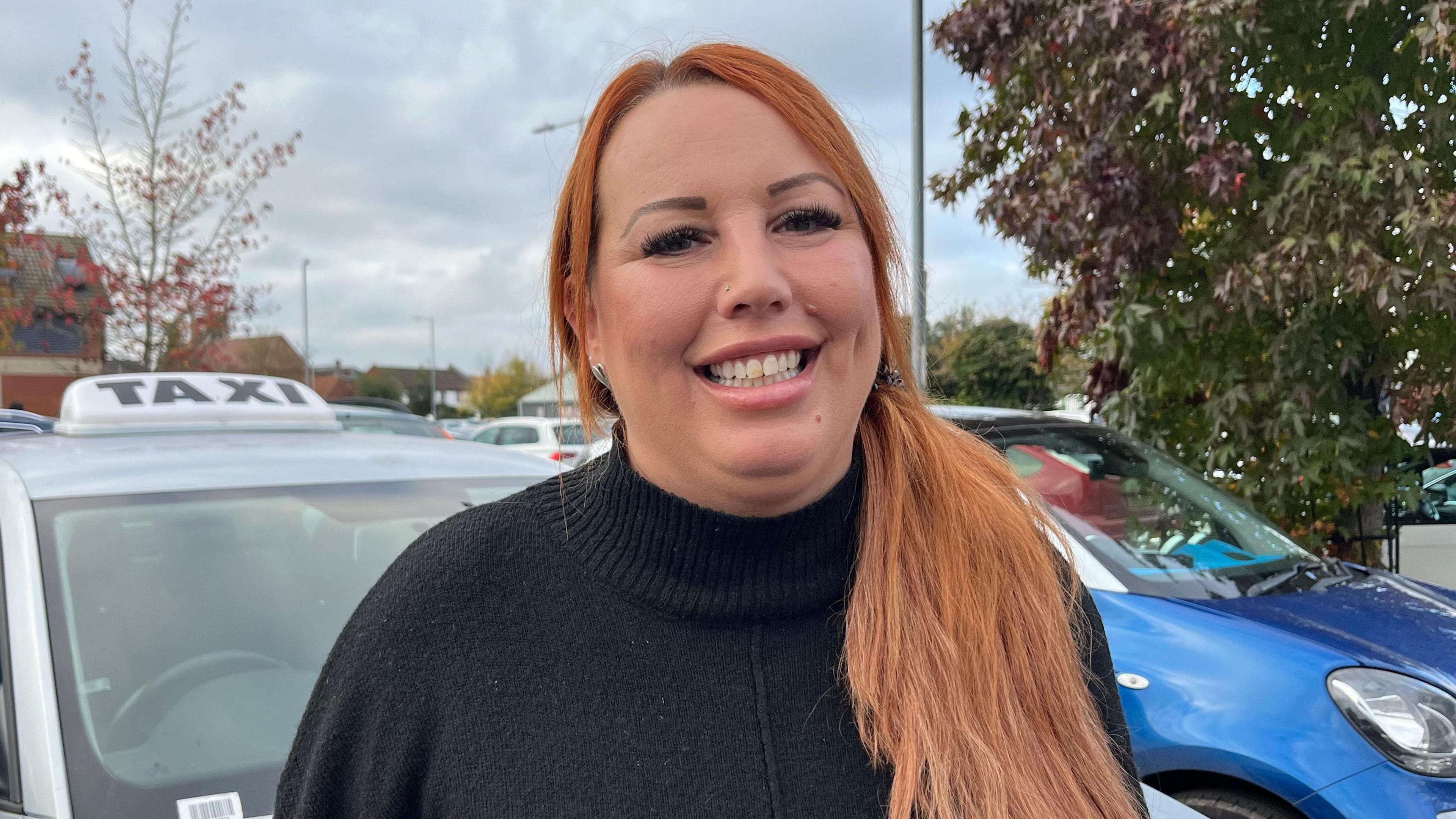 Jenny Wilson smiles at the camera as she is photographed outside, in front of a car. She is wearing a black roll-neck jumper and has long auburn hair. 