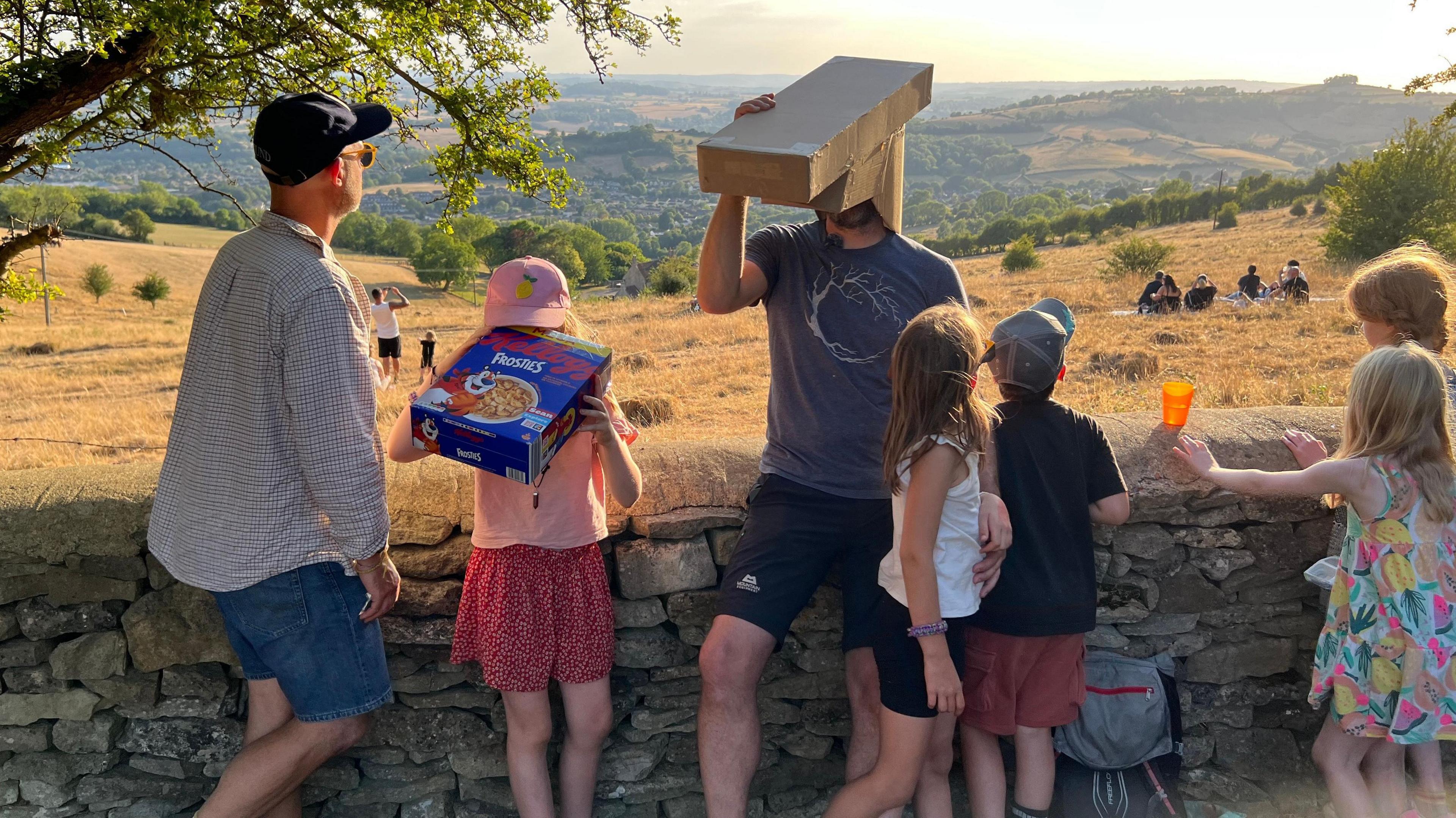 People are stood by a stone wall looking through cardboard boxes up at the eclipse