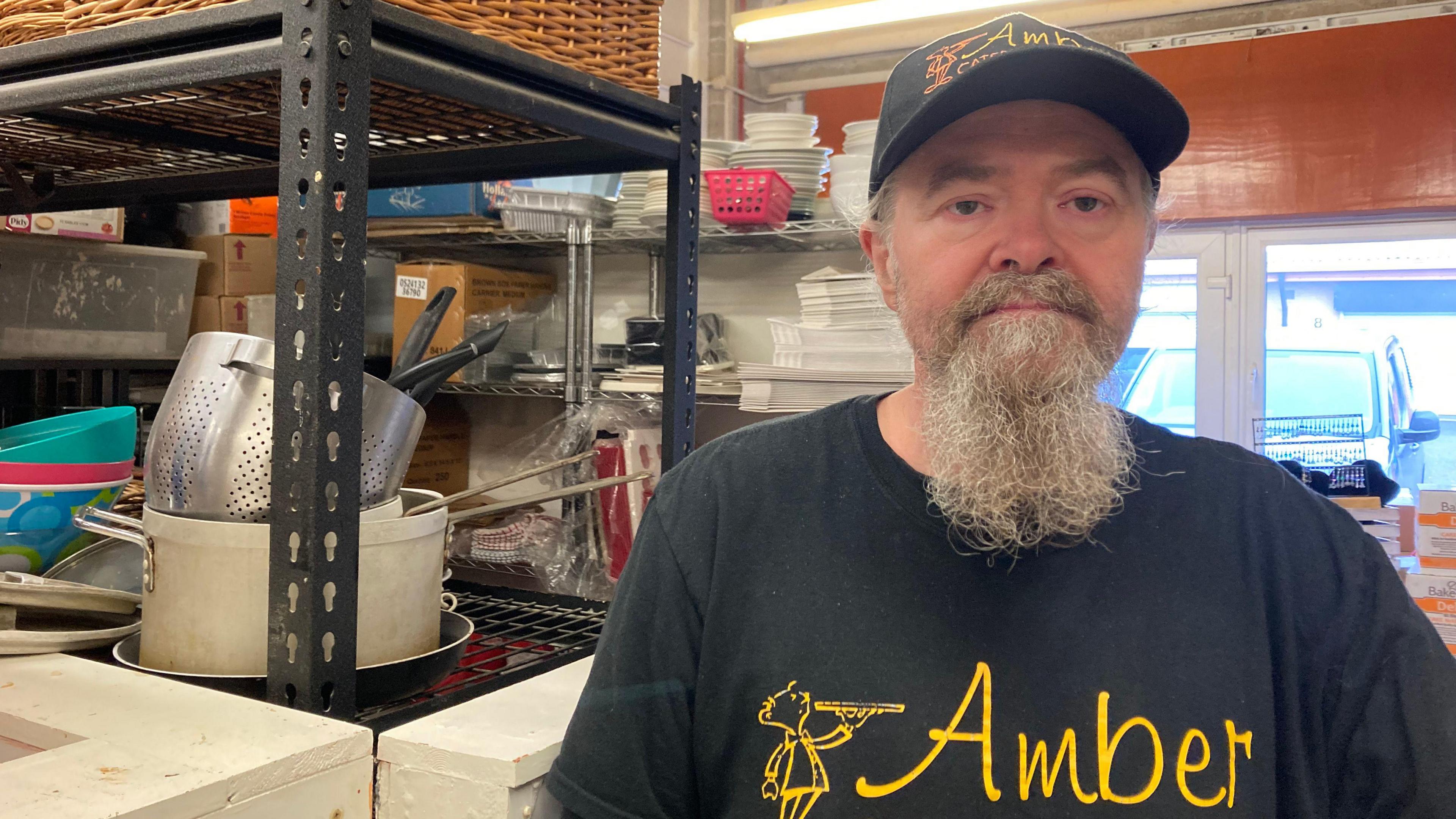 A man, standing to the right of the photo, looking at the camera. He has a grey beard. He is wearing a black hat and a black t-shirt with the word "Amber" written in yellow on it. There is shelving to the left of him, with pots and pans stacked up on it. 