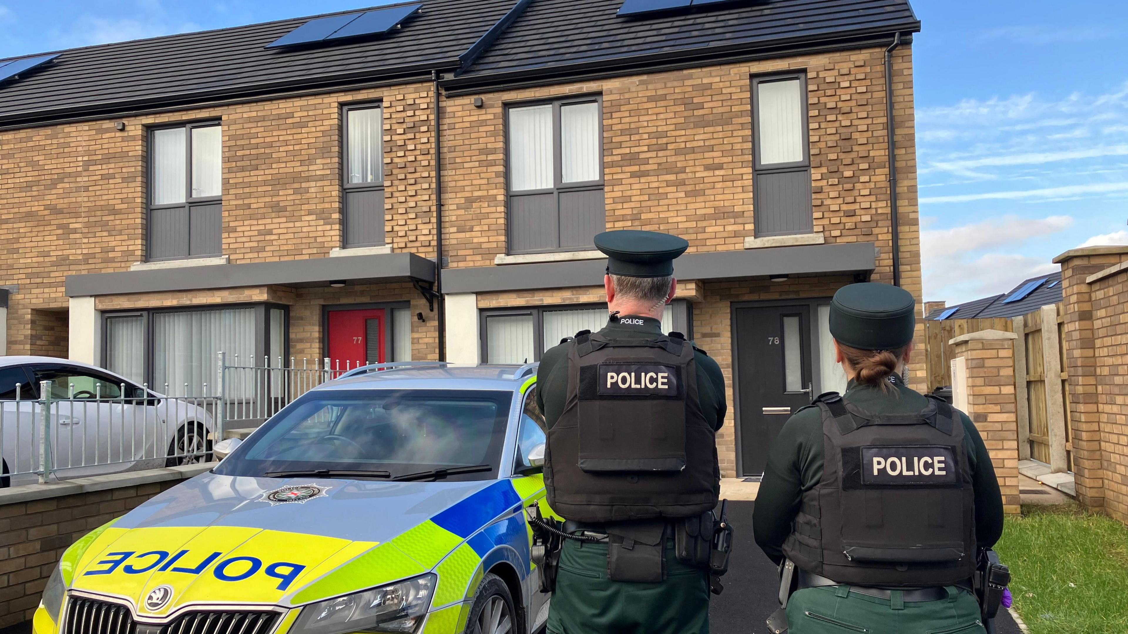 A still image of two police officers with their backs turned to the camera outside a light brick terrace house. There is a police car parked in the driveway.