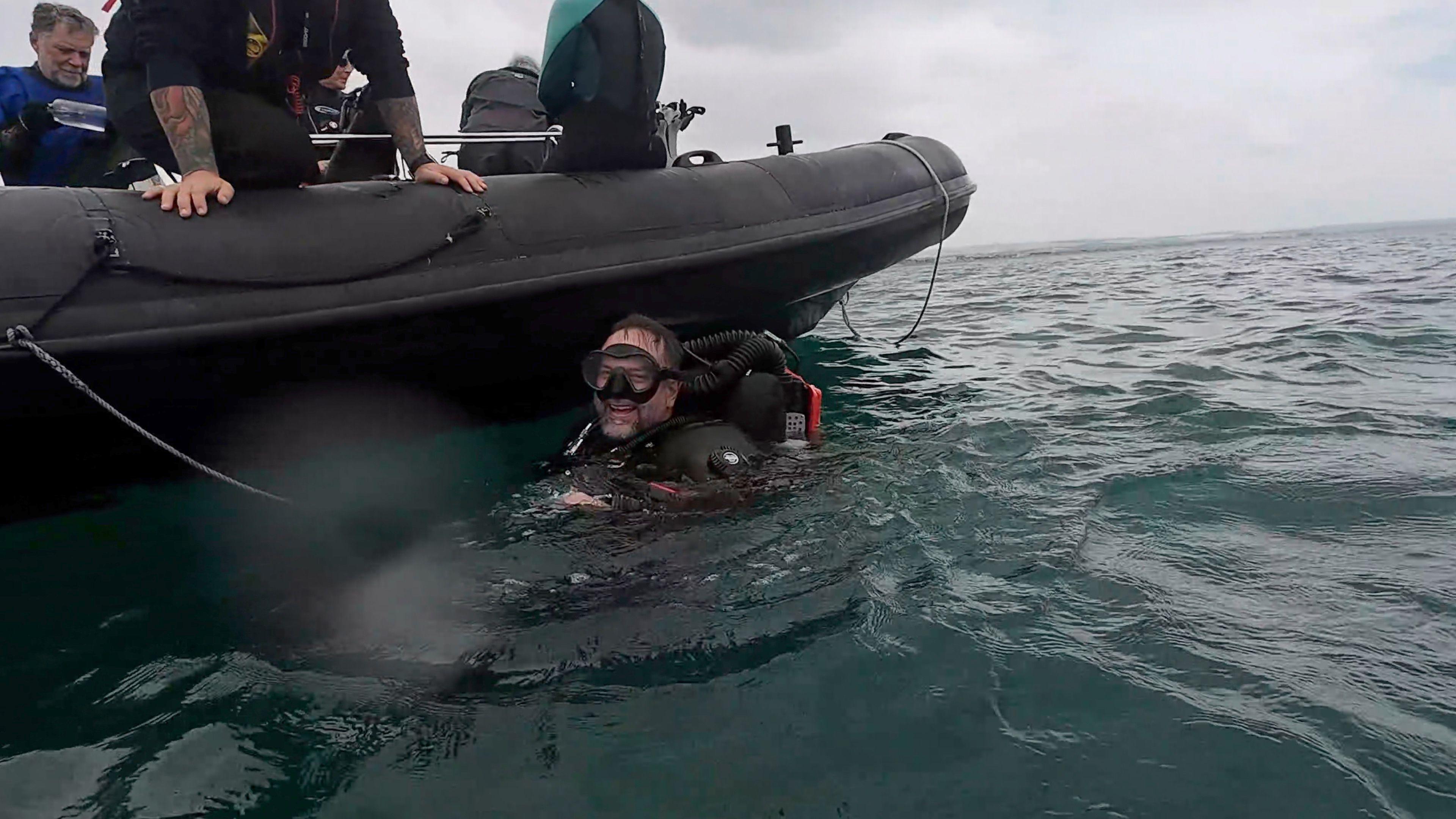A man in a diving suit smiles as he bobs in the sea by a power boat.