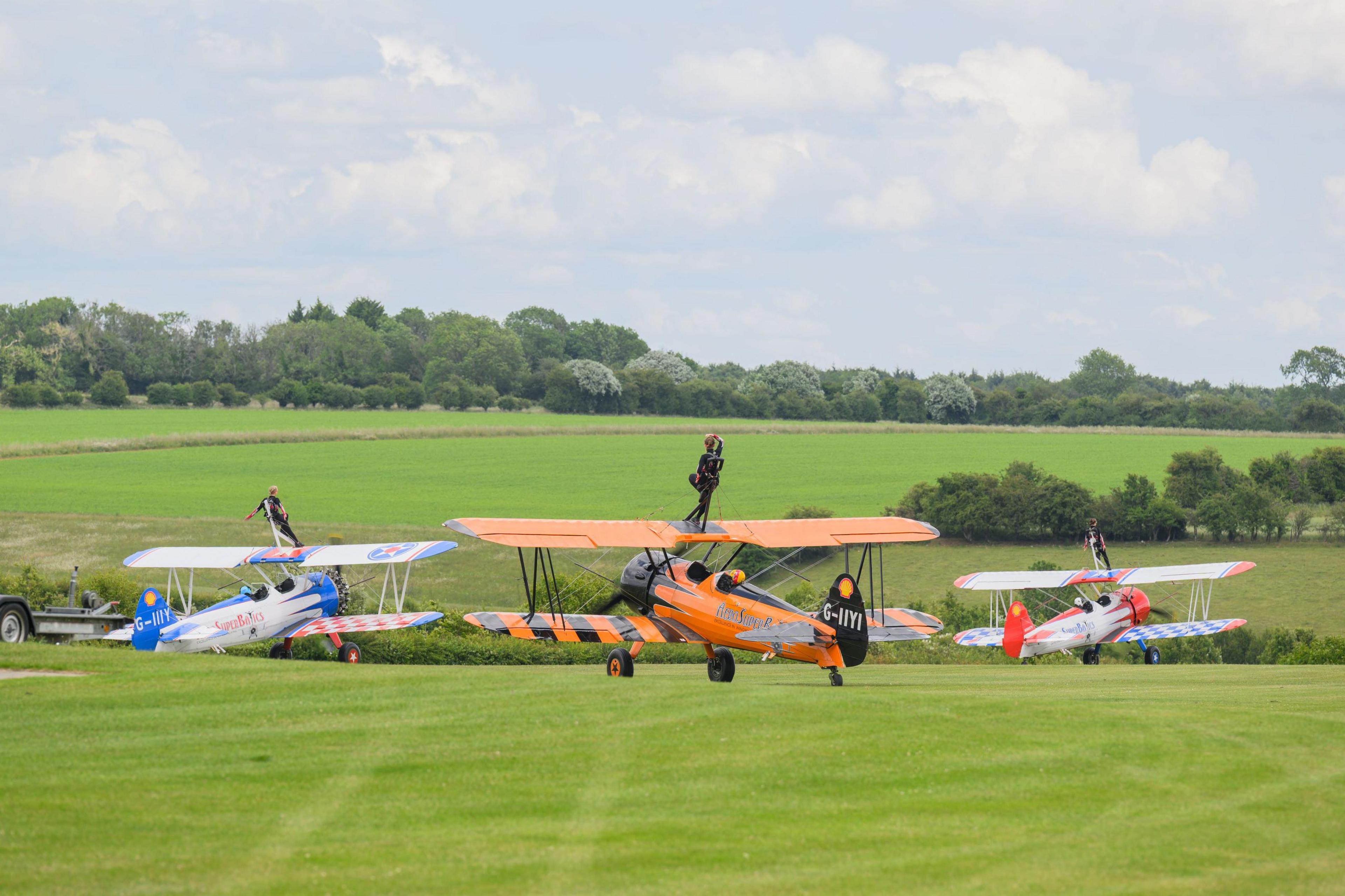 Three bi-planes coloured white blue and red, orange and black and red and white, in an airfield with three female wing walkers on the top deck of each aircraft.