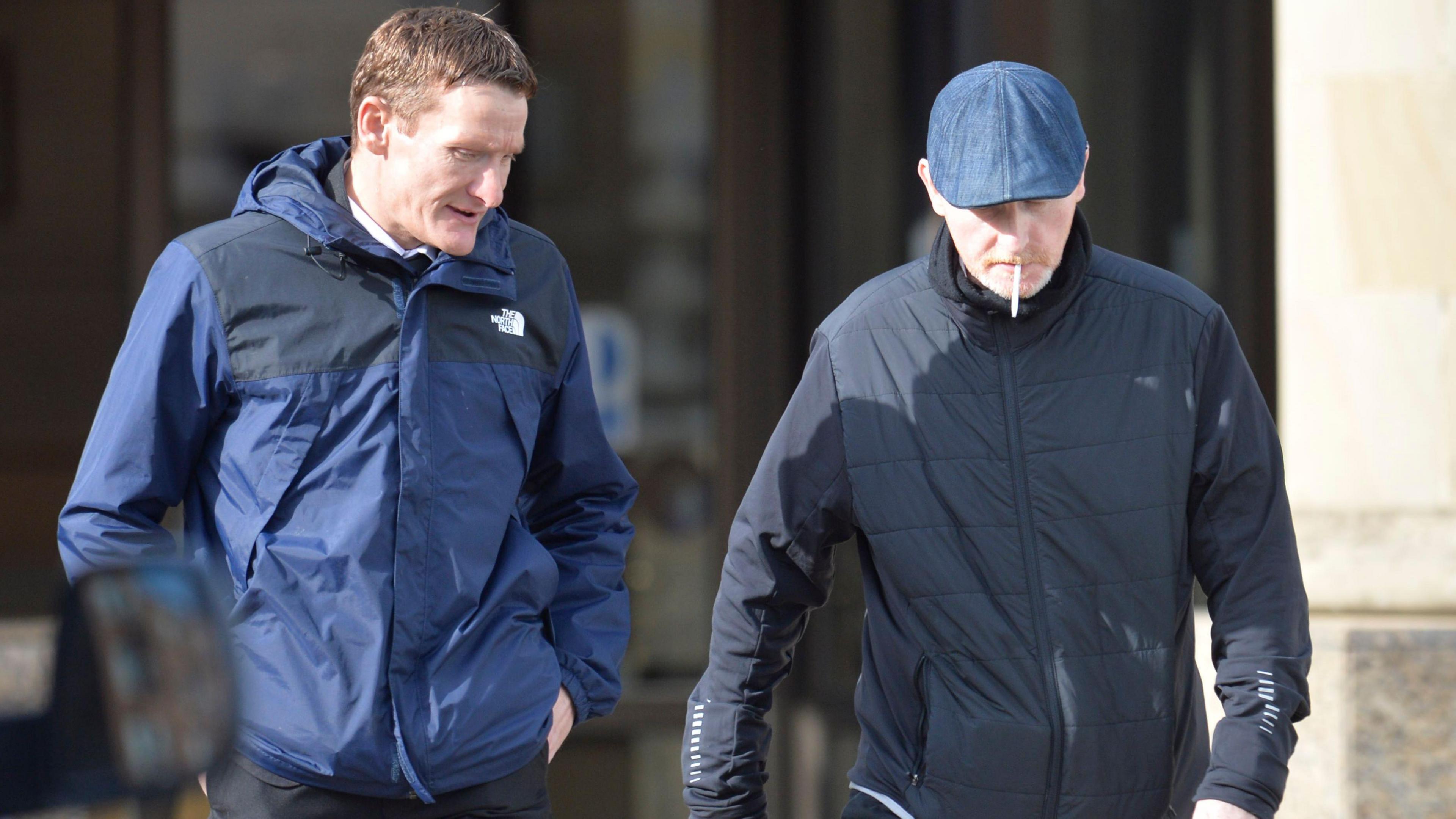 John Clark and Barry Watson pictured walking while in conversation outside the High Court in Glasgow. Clark, who has short red hair, is looking down to his left. He is wearing a blue North Face hooded jacket. Watson is looking down at the pavement and has a cigarette hanging from his mouth. He is wearing a quilted navy jacket and a navy skip cap. 