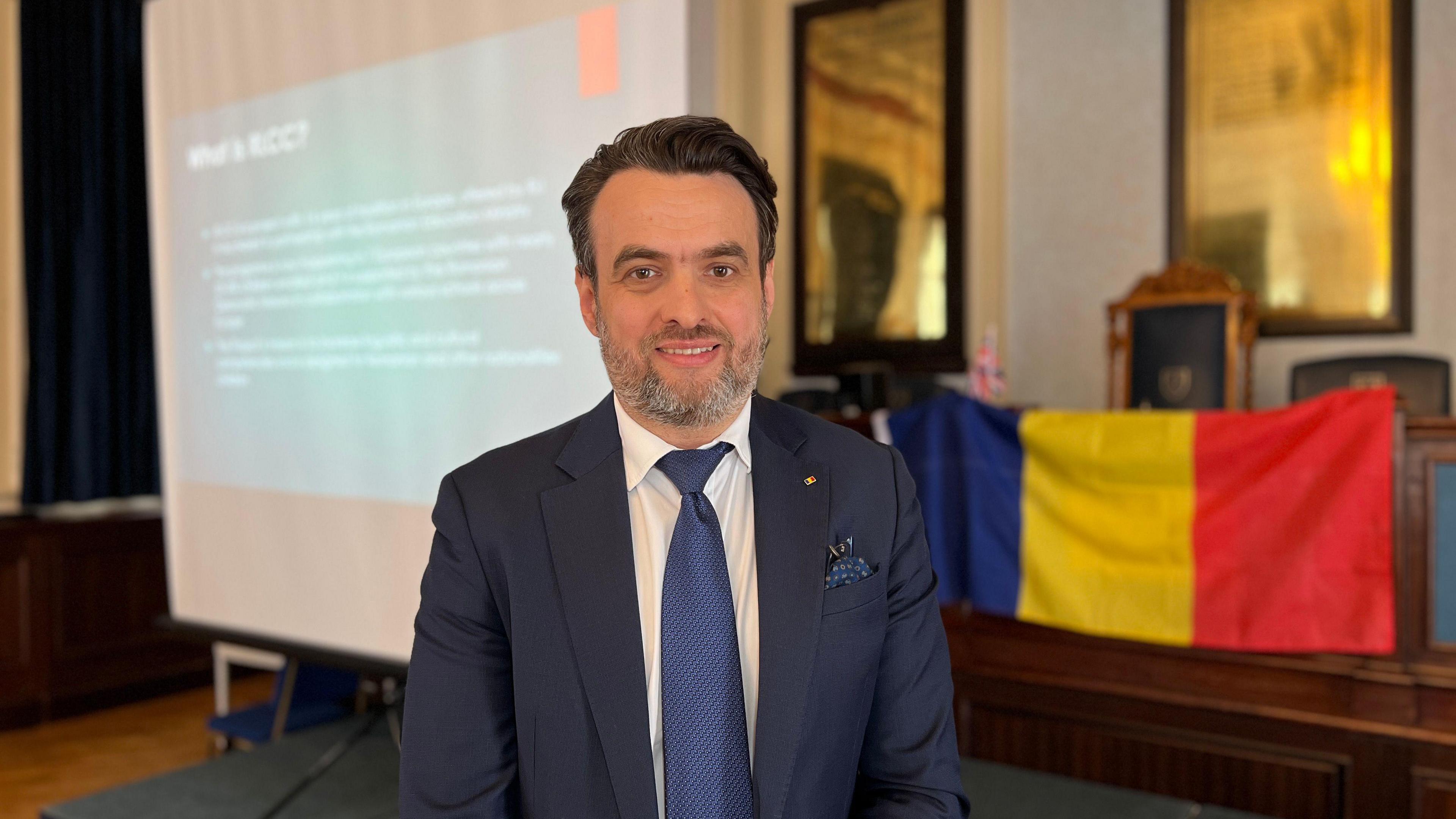 Robert smiles at the camera as a projector screen show the details of the course and there's a Romanian flag next to it