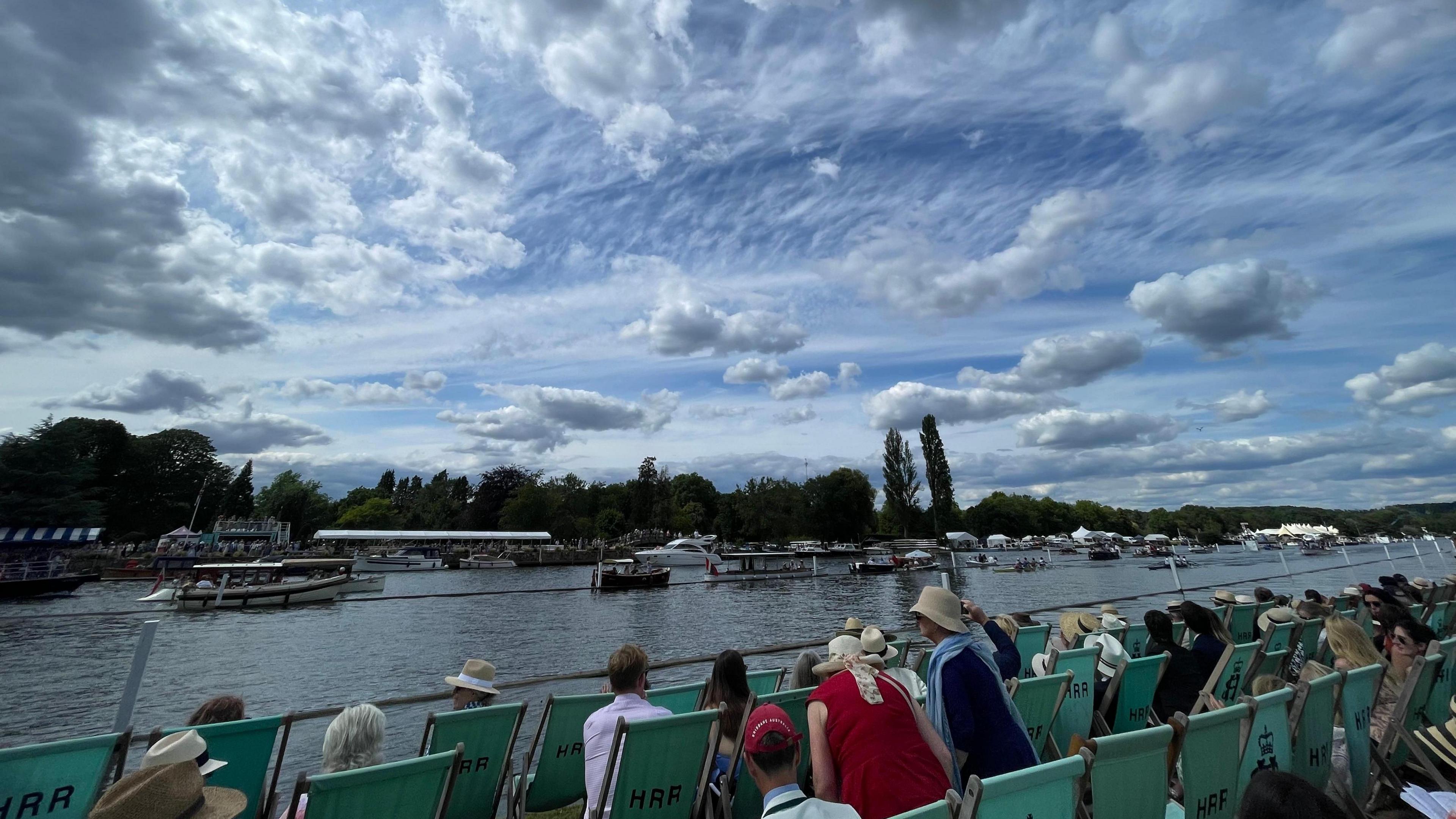Cloudy skies hang above the River Thames at Henley as members of the regatta take to teal coloured deckchairs on the banks of the rover to watch the rowing races and small boats go past.
