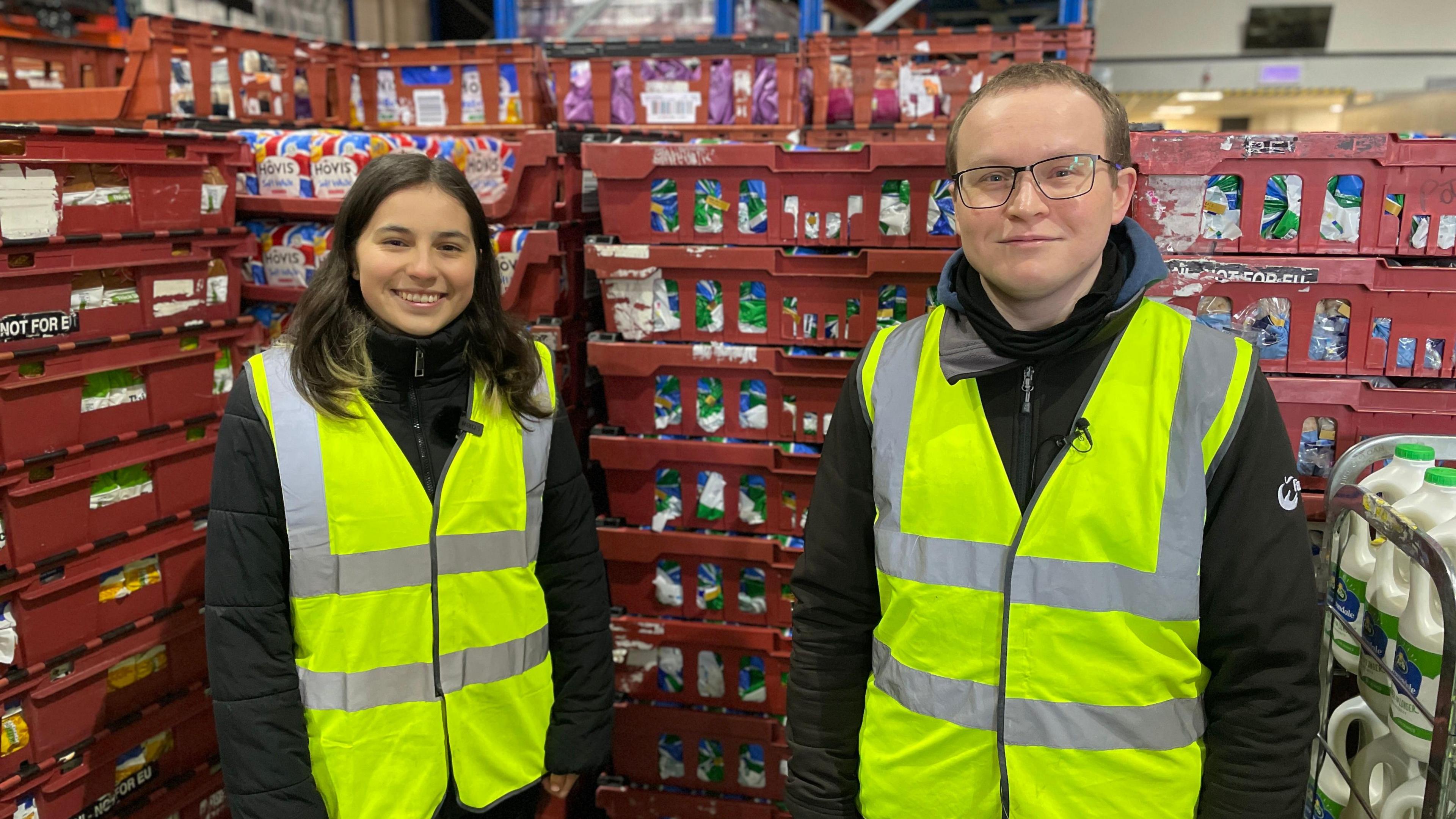 Isabella stands next to Jonathan Williams, a young man with short hair and young glasses, who is also a wearing a luminous jacket. They are both standing in front of shelves packed with food.