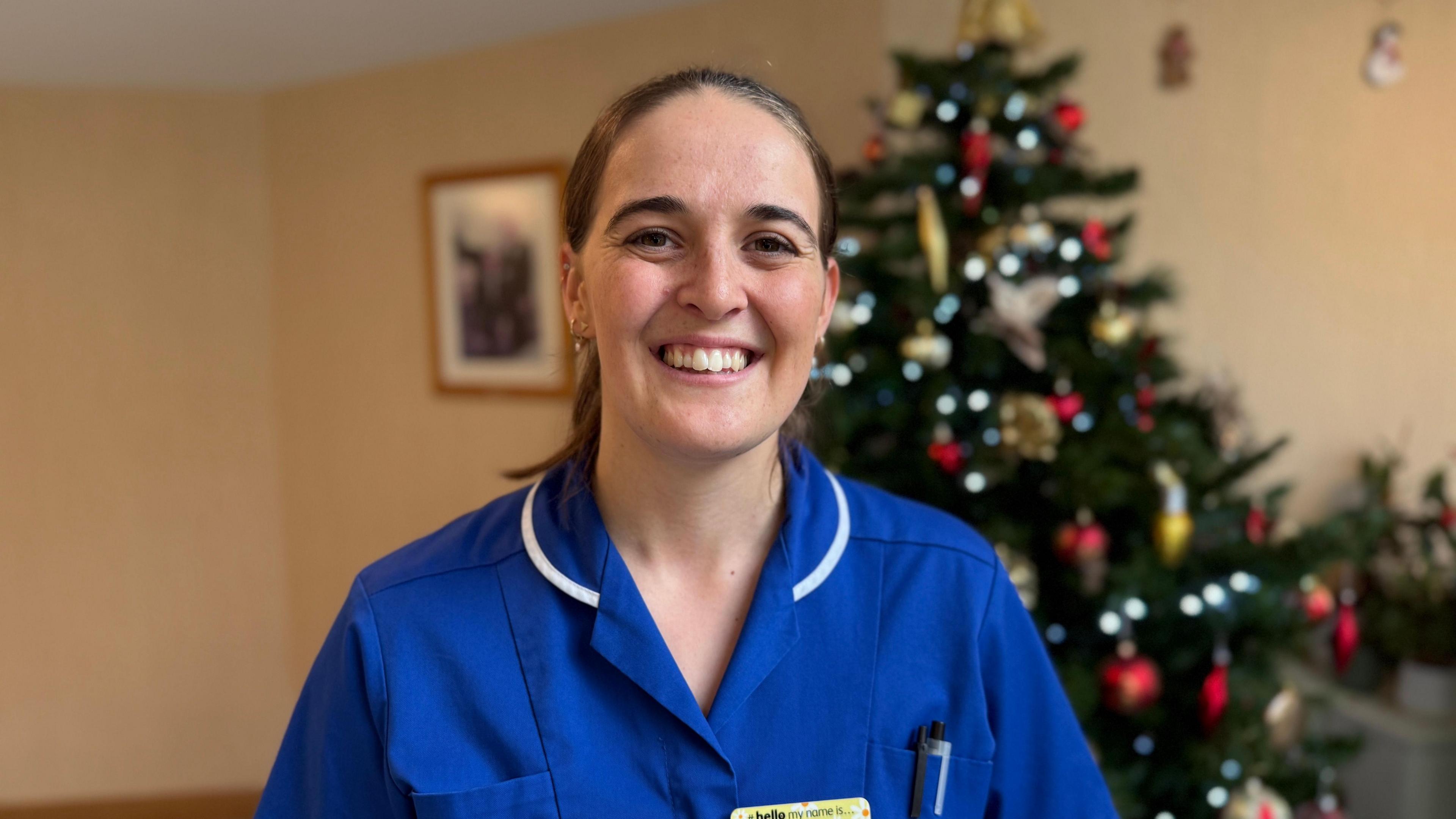 A woman wearing a bright blue nurse uniform looking towards the camera and smiling. She has brunette hair and is standing in front of a Christmas tree.