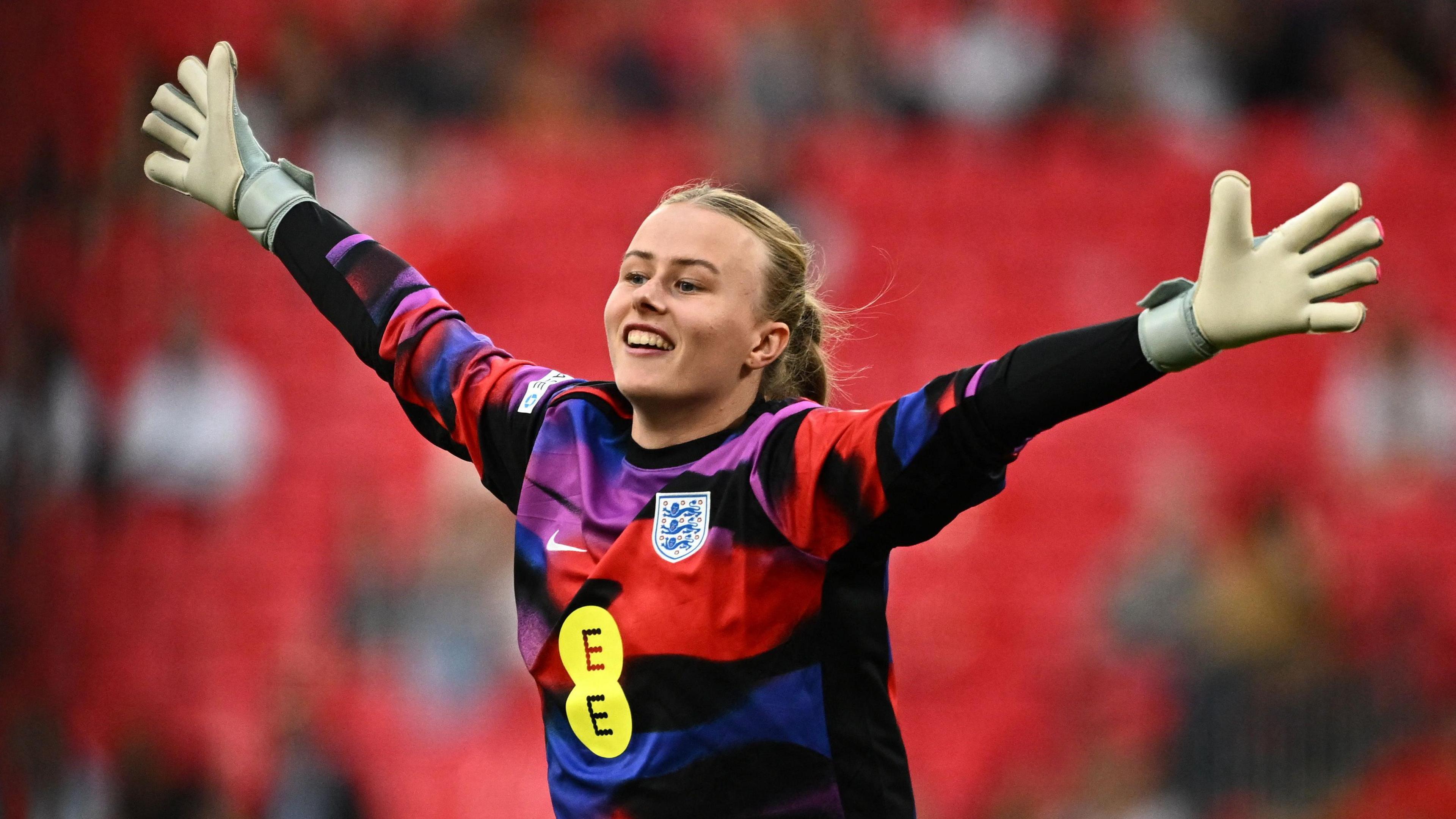 A woman with a football shirt and hands in the air celebrating. She wears goalkeeper gloves and has blonde hair, pulled back in a ponytail