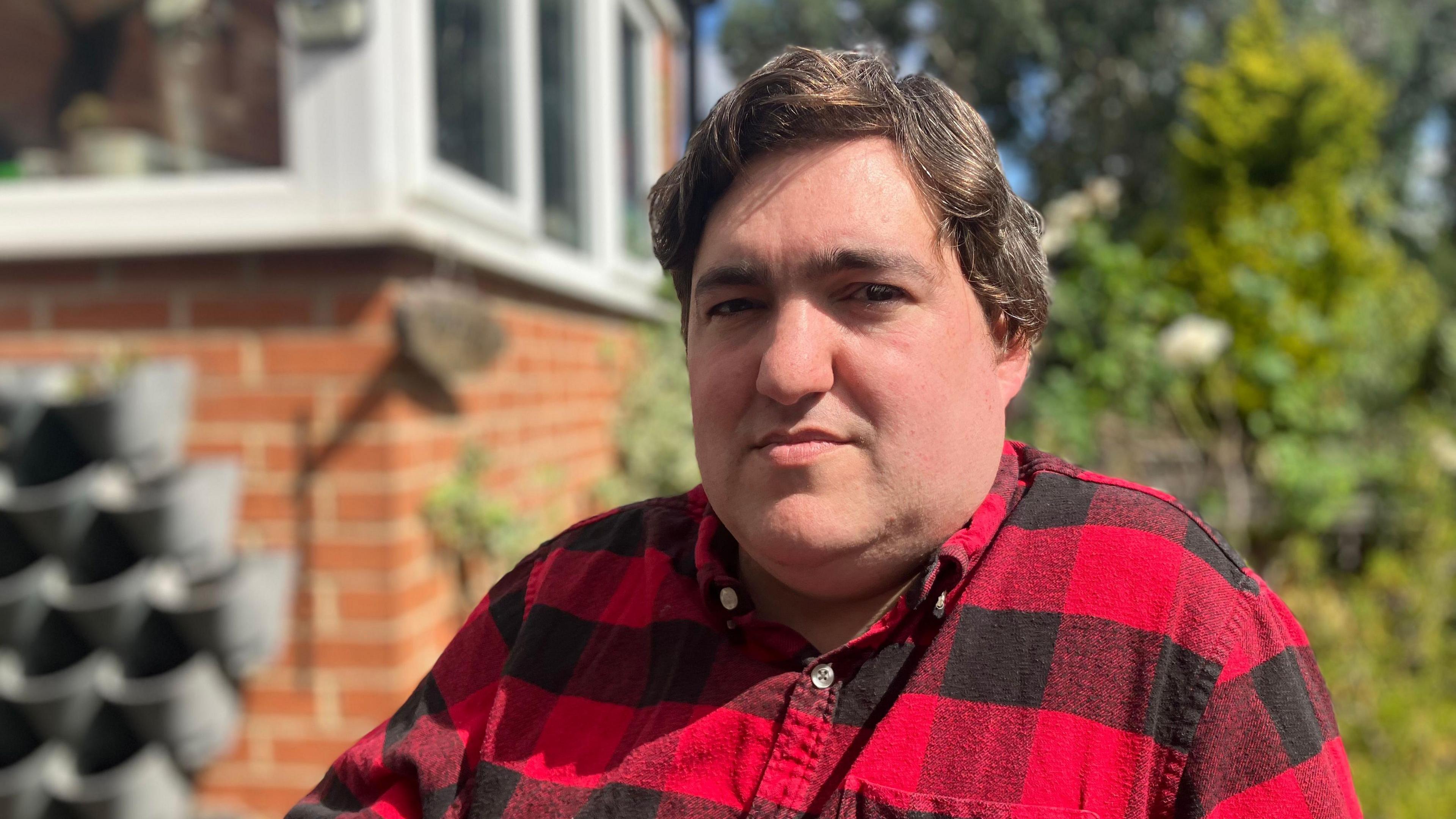 A man with a red and black shirt looks at the camera. Behind him a bush is blurred alongside a brick wall.