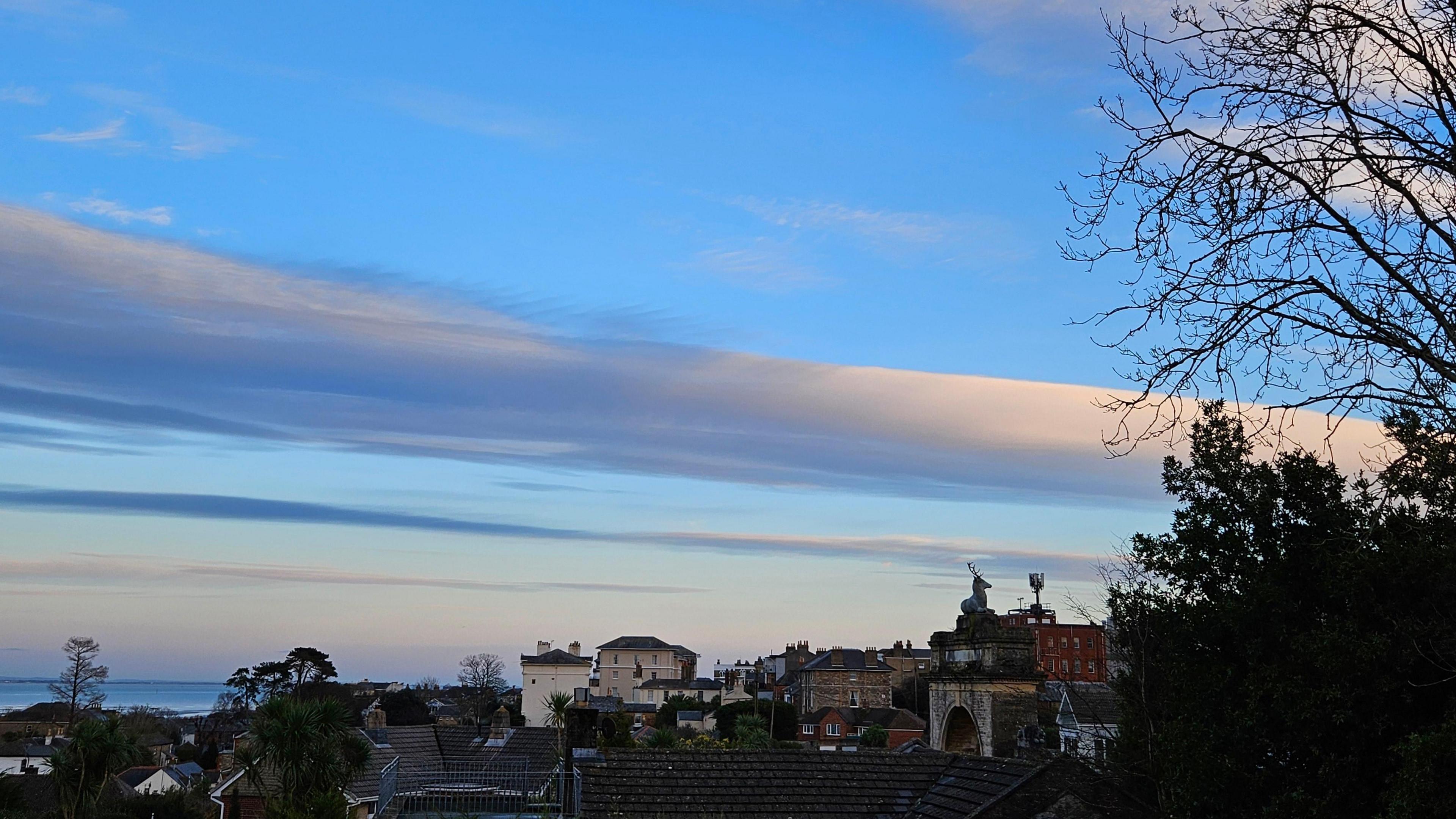 A blue sky, with white clouds. The clouds are lit up from the setting sun. At the bottom of the photo you can see roof tops of various homes, including a large stag on top of an arch. 