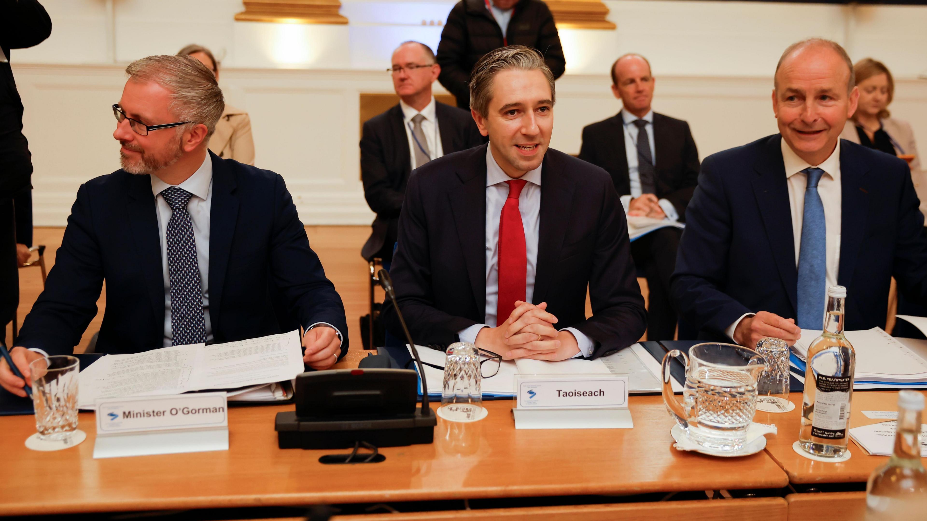 Three men - Roderic O'Gorman, Simon Harris and Micheál Martin - sit together at a large table. On the left, O'Gorman is wearing a suit and tie. He has grey hair, is wearing rectangle shaped glasses and is looking to the side. Harris sits in the middle and is mid-speech, wearing a suit and tie. Martin is sat on the right and smiles while wearing a suit and tie.