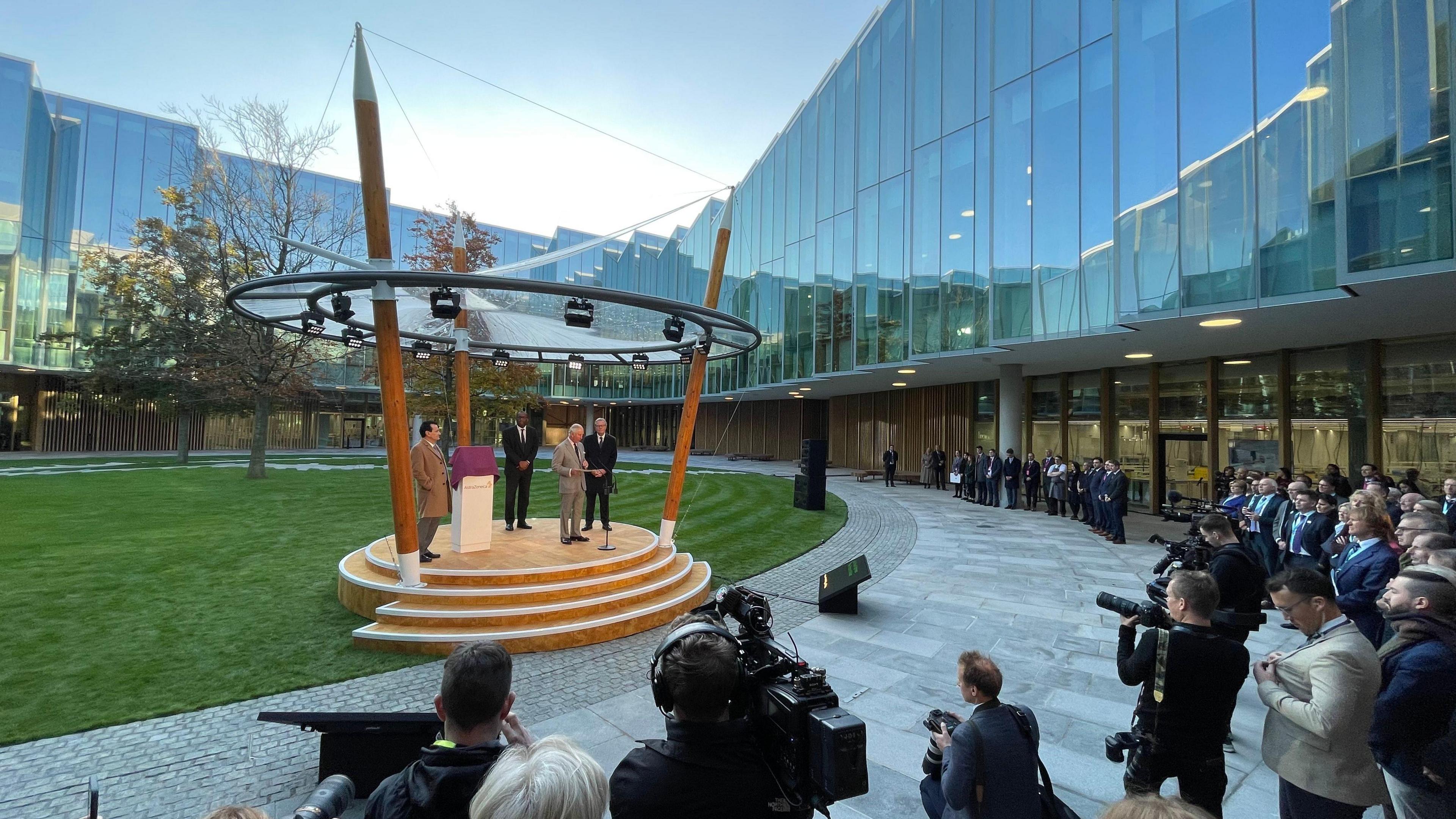 Slightly to the left of the centre of frame is a small stage, with four steps leading up to a wooden platform. The stage has three large wooden spears coming out of it, which are holding up a roof and a lighting rig. There are four people standing on the stage, among them is Prince Charles, as he was at the time. The stage is standing on a well-kept, freshly mown, green lawn, which appears to be oval in shape. The lawn is inside the middle of a large, glass fronted, angular building. A crowd of people, including photographers and camera crews, is watching what is happening on the stage.