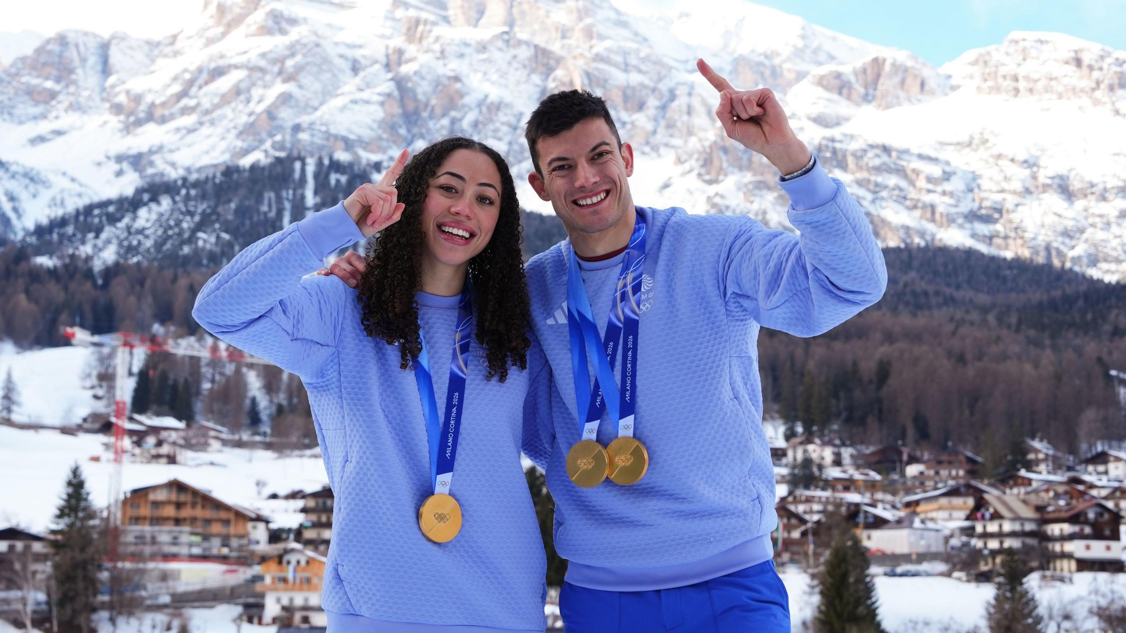 Tabby Stoecker (left) and Matt Weston stand with one arm around each other and the other in the air pointing their index fingers. They are both wearing Team GB kit and Winter Olympic gold medals around their necks. There are snow-capped moutains in the background