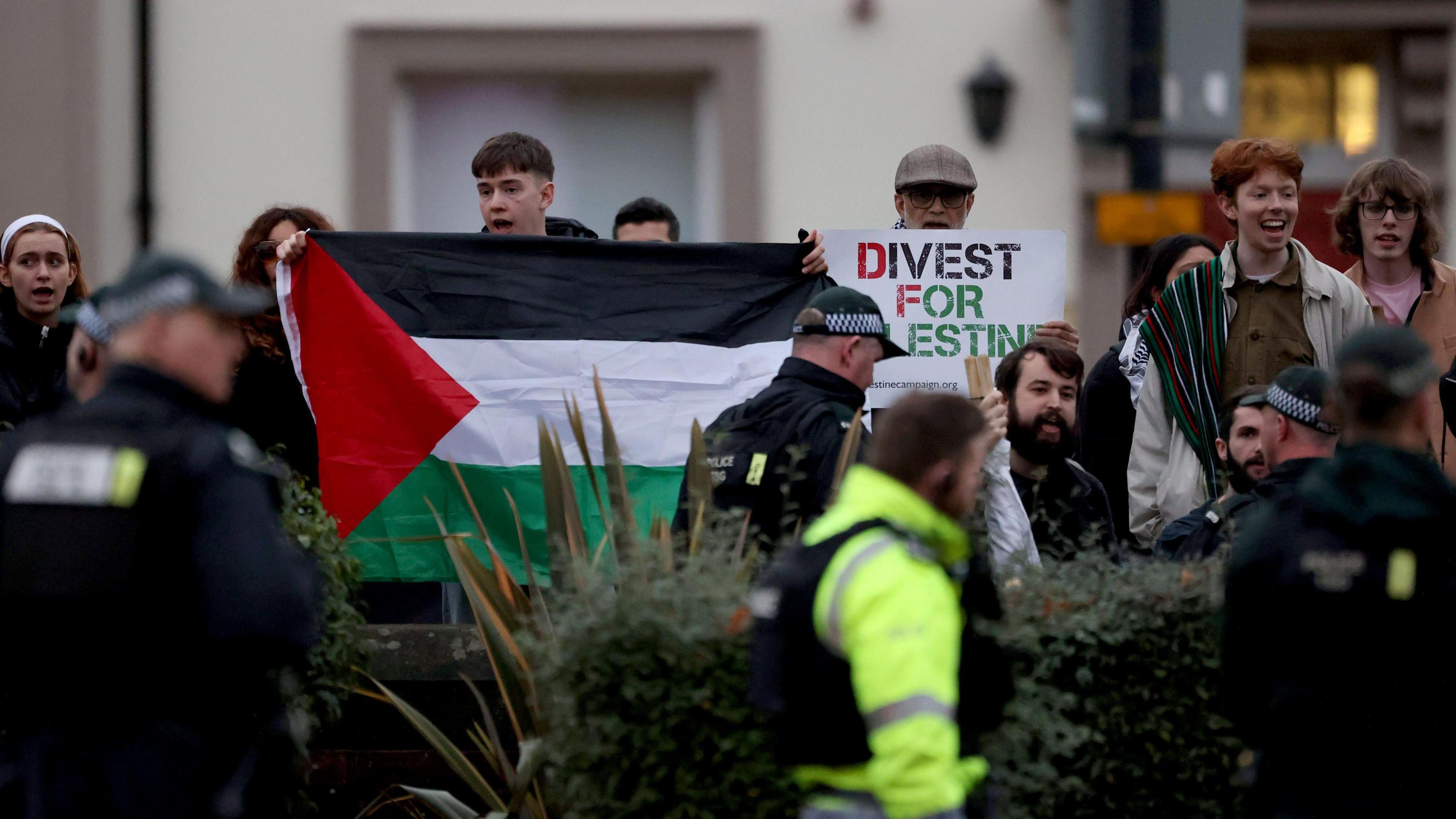 A group of pro-Palestinian protestors standing behind a wall and hedge holding a Palestinian flag and signs. Police officers are seen on patrol and  engaging with protestors.