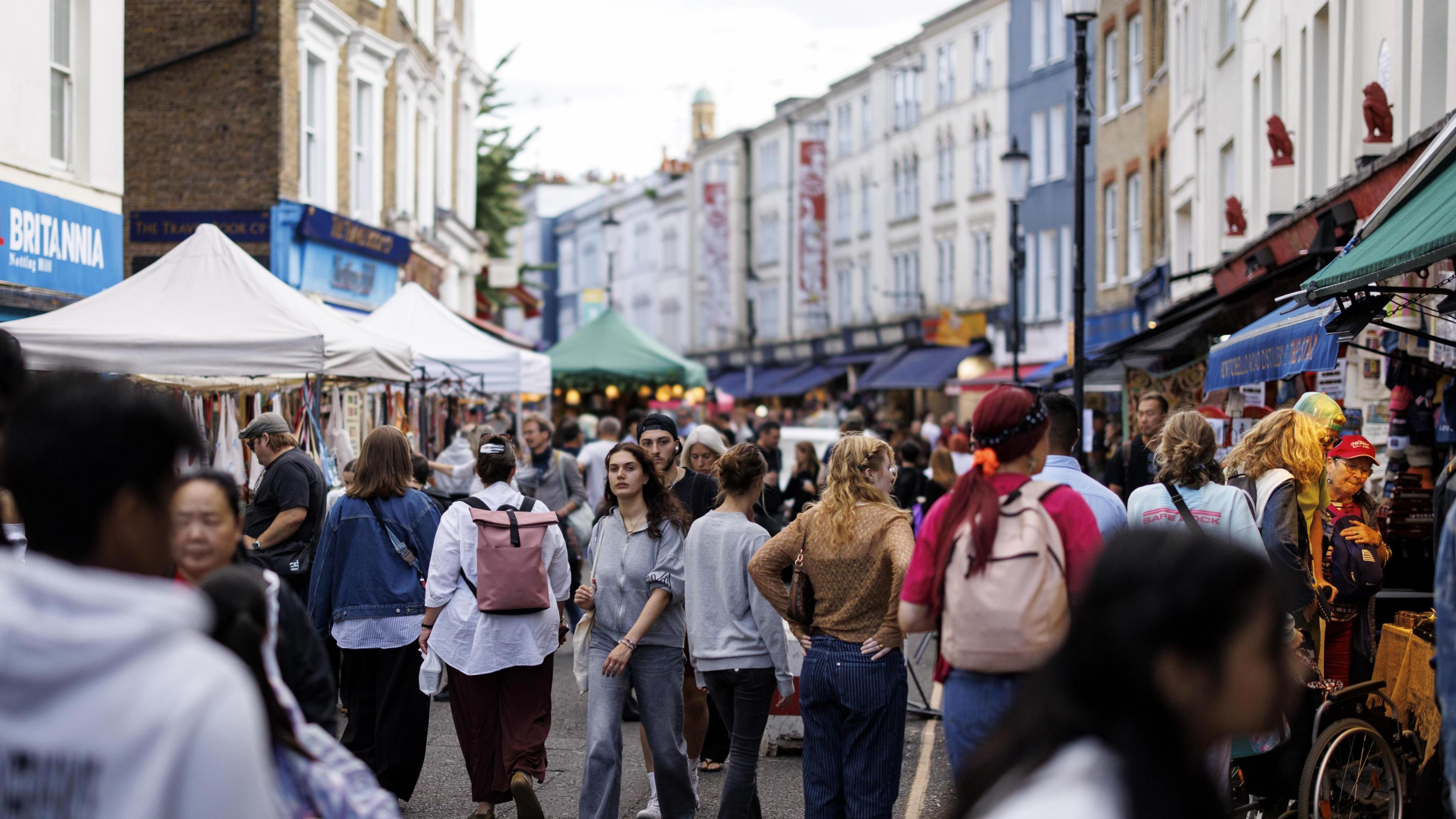 A street with stalls on both sides of the road with many people milling about. There are tall houses visible in the background.