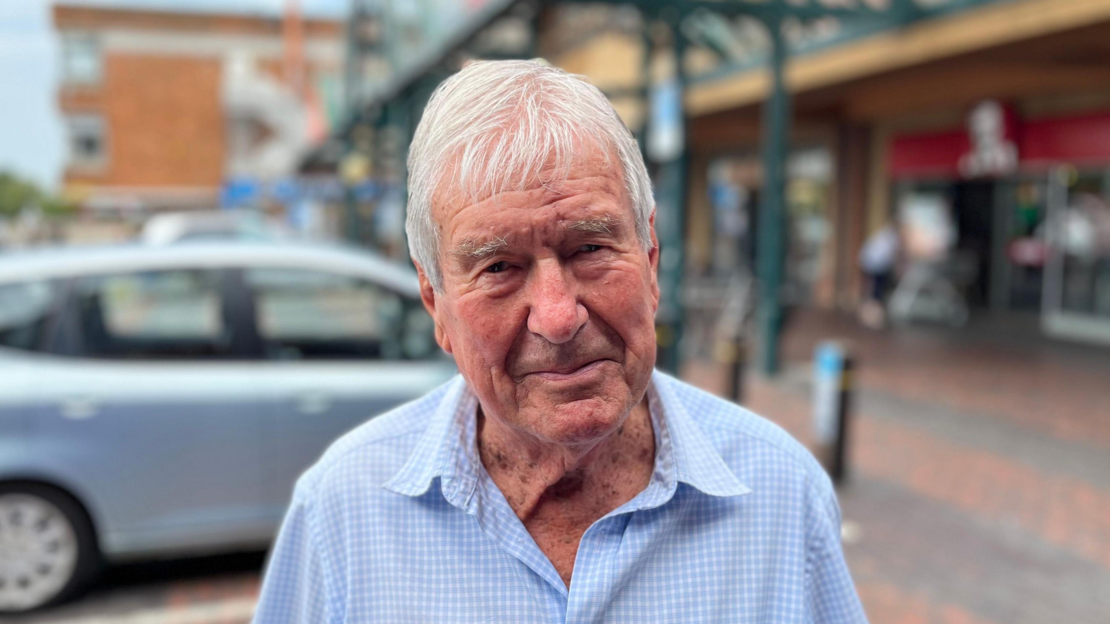 Man looking directly at the camera with a plain expression. He is wearing a blue and white gingham shirts. The background is blurred but he is stood in front of a blue car in a car park 