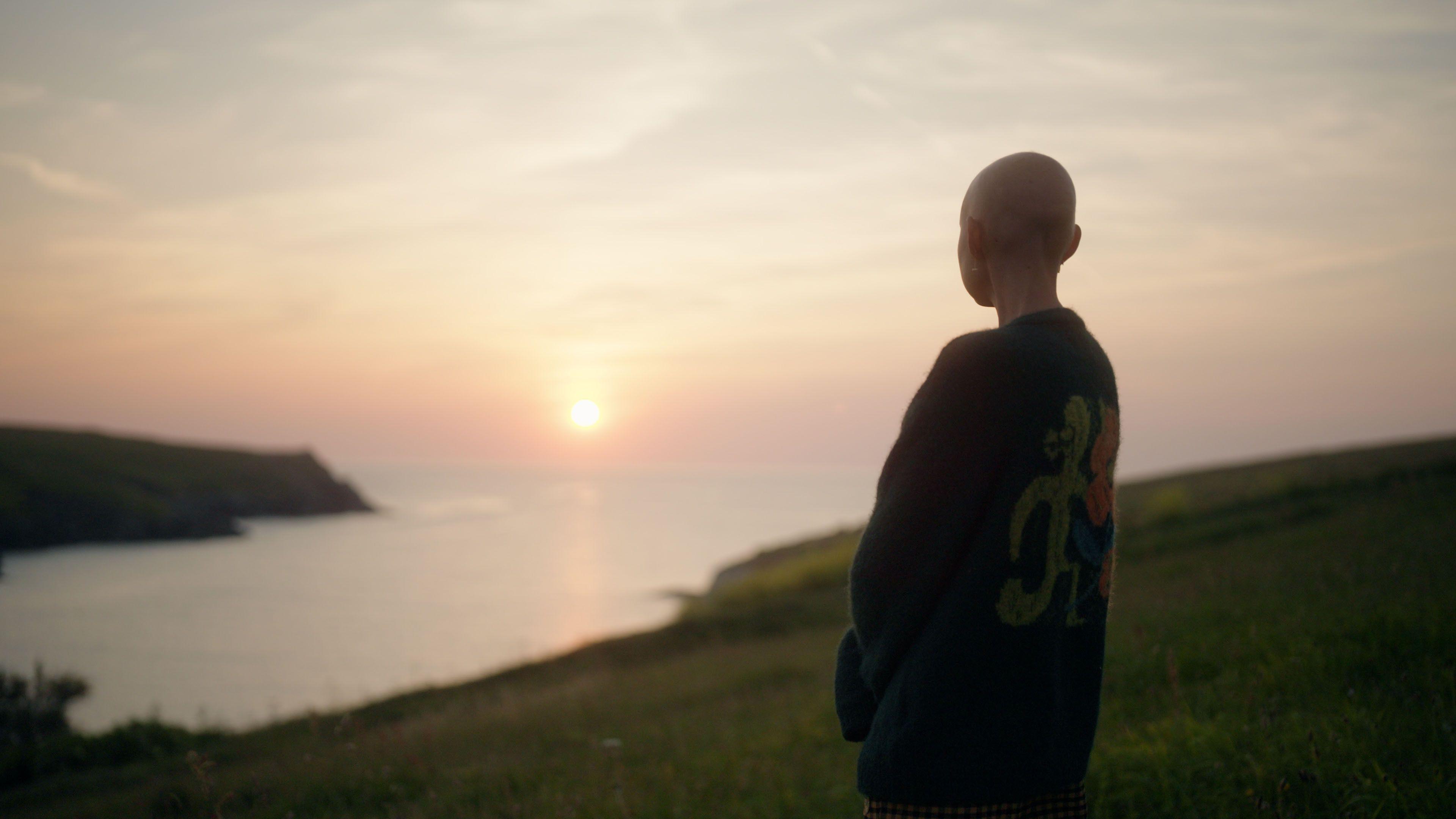 Kris is standing on the coastline. She is wearing a cardigan and is looking out on to the horizon. The Sun can be seen shining in the distance.