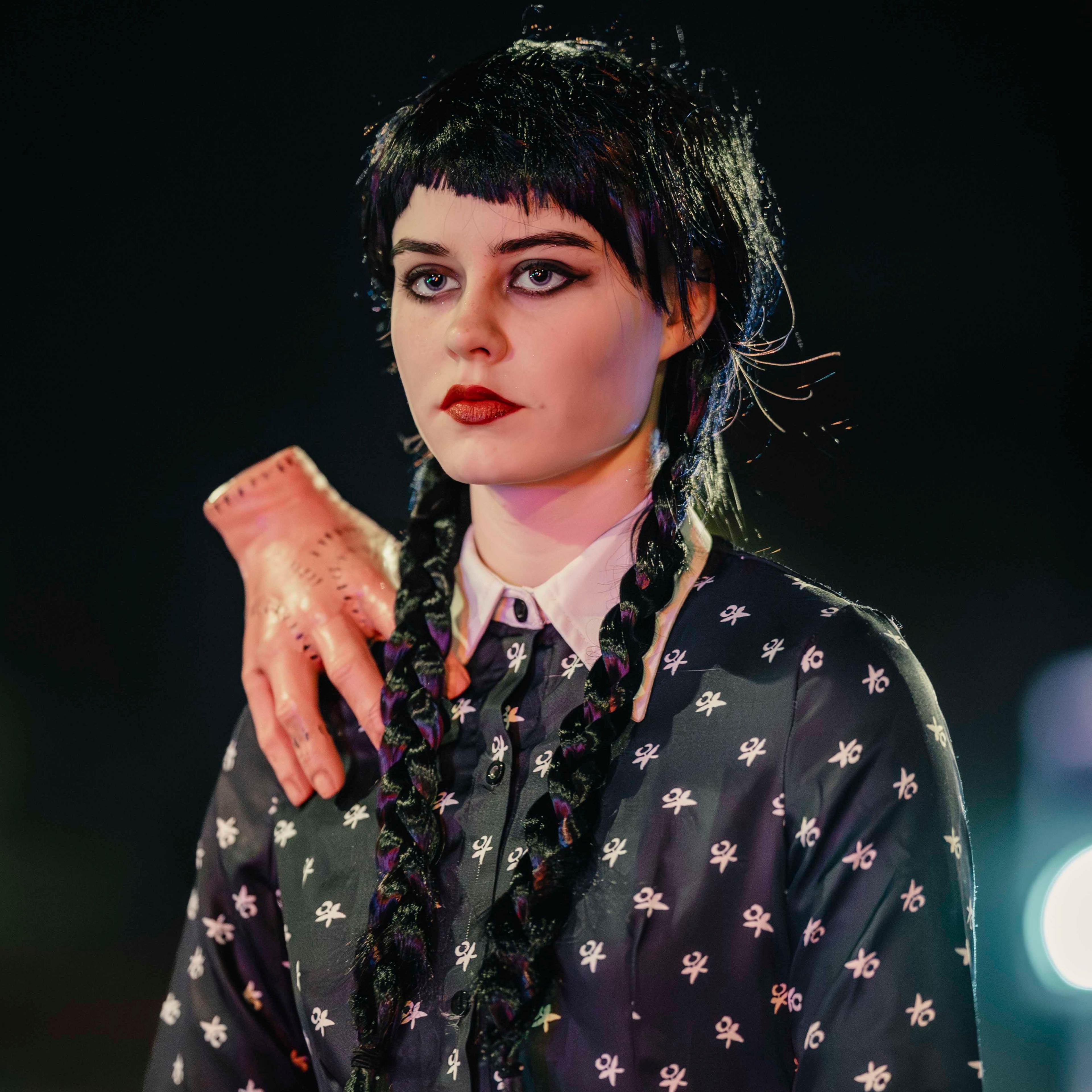 A young woman with long black hair in two plaits wearing a black and white dress, buttoned at the neck. She has a disembodied hand resting on her left shoulder.