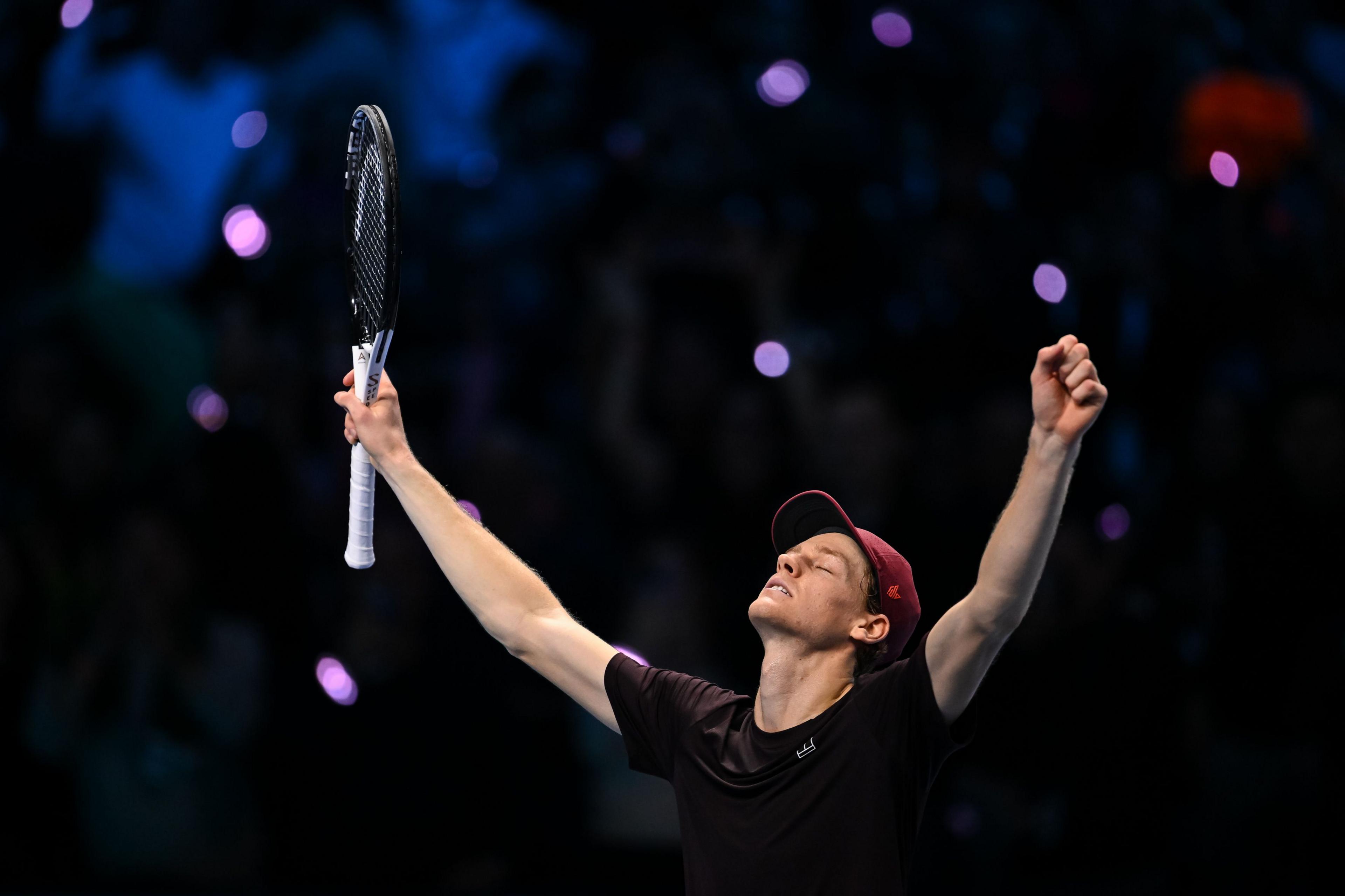 Tennis player raising both arms and holding a racket in celebration on a dark court, with blurred audience and bright lights in the background.