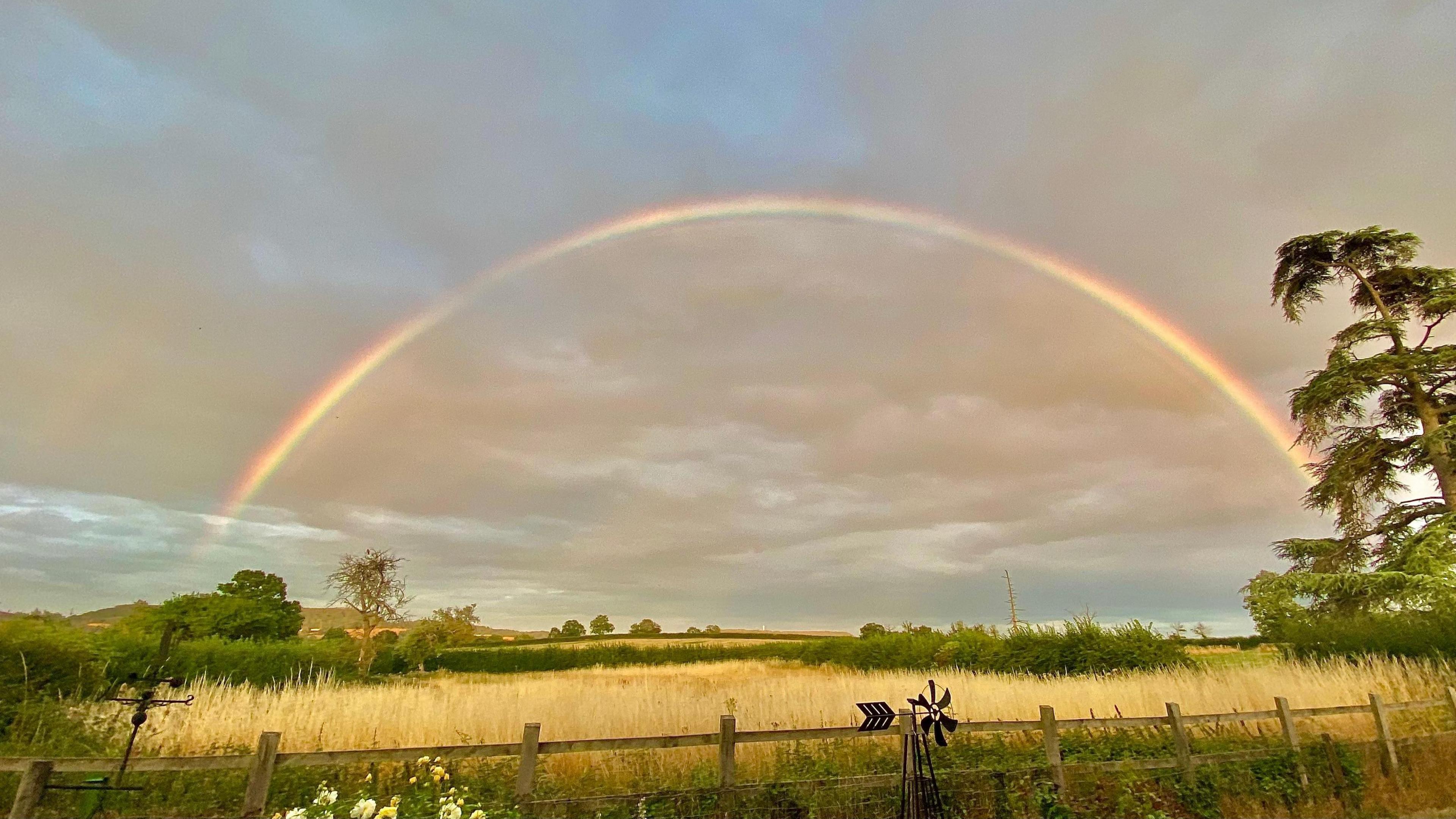 Rainbow across a field with a grey sky in the background. Trees and hedgerows are also visible and there are grey clouds in the sky