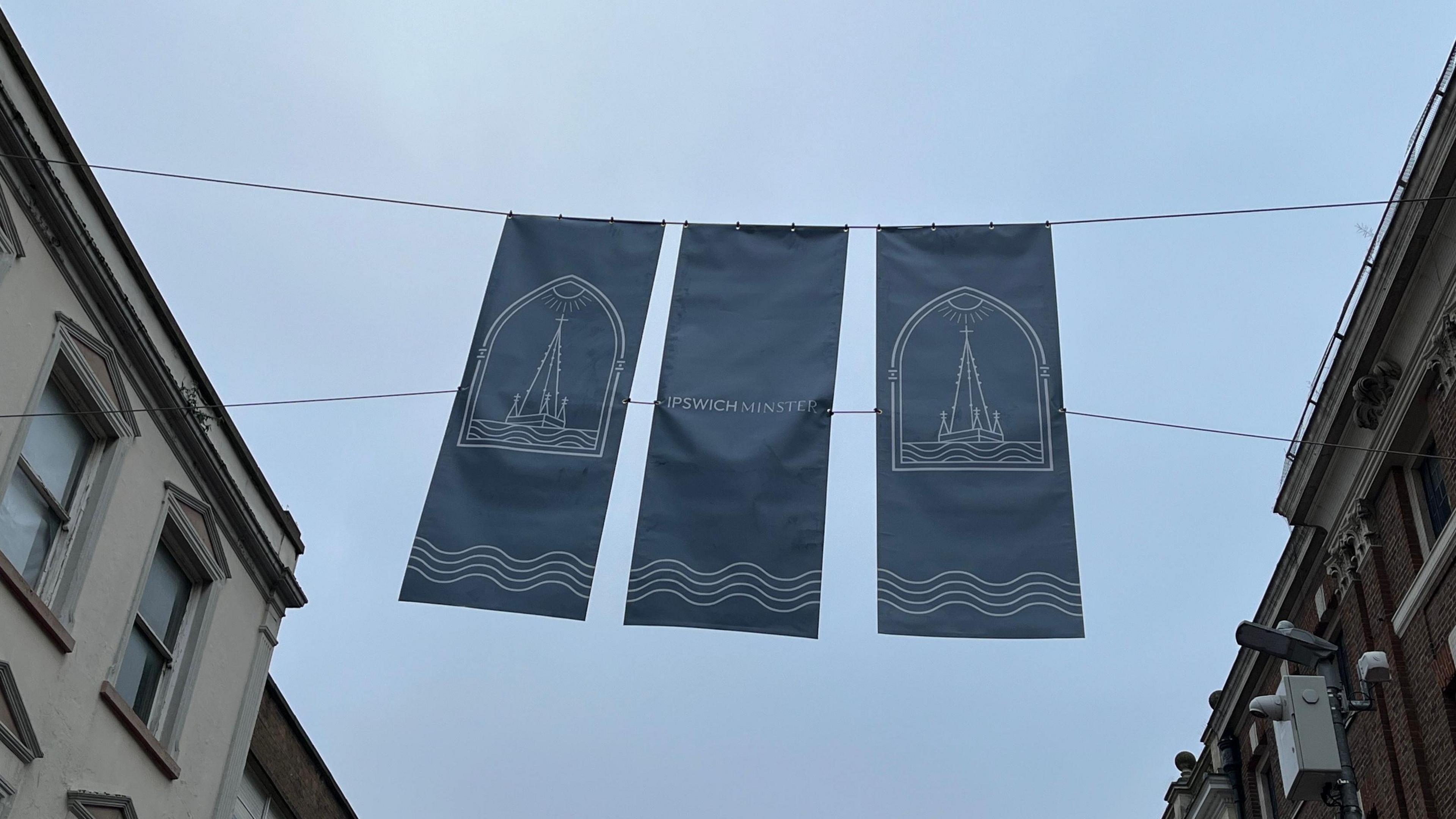 Three navy blue banners hanging portrait above a pedestrian street. They feature illustrated images of the tower and the words "Ipswich Minster".